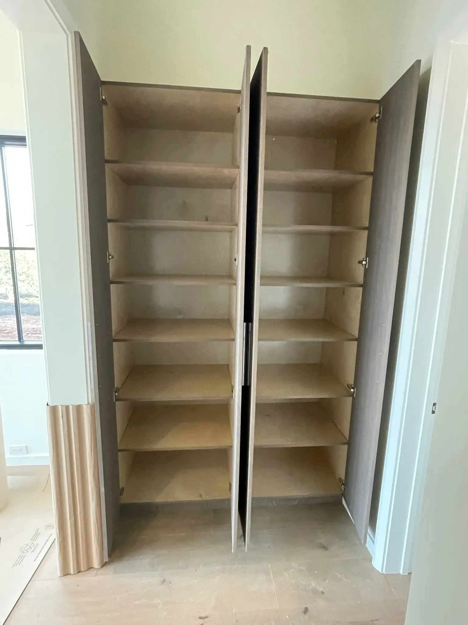 Open closet with empty wooden shelves and a central divider, doors open in a room with a window.
