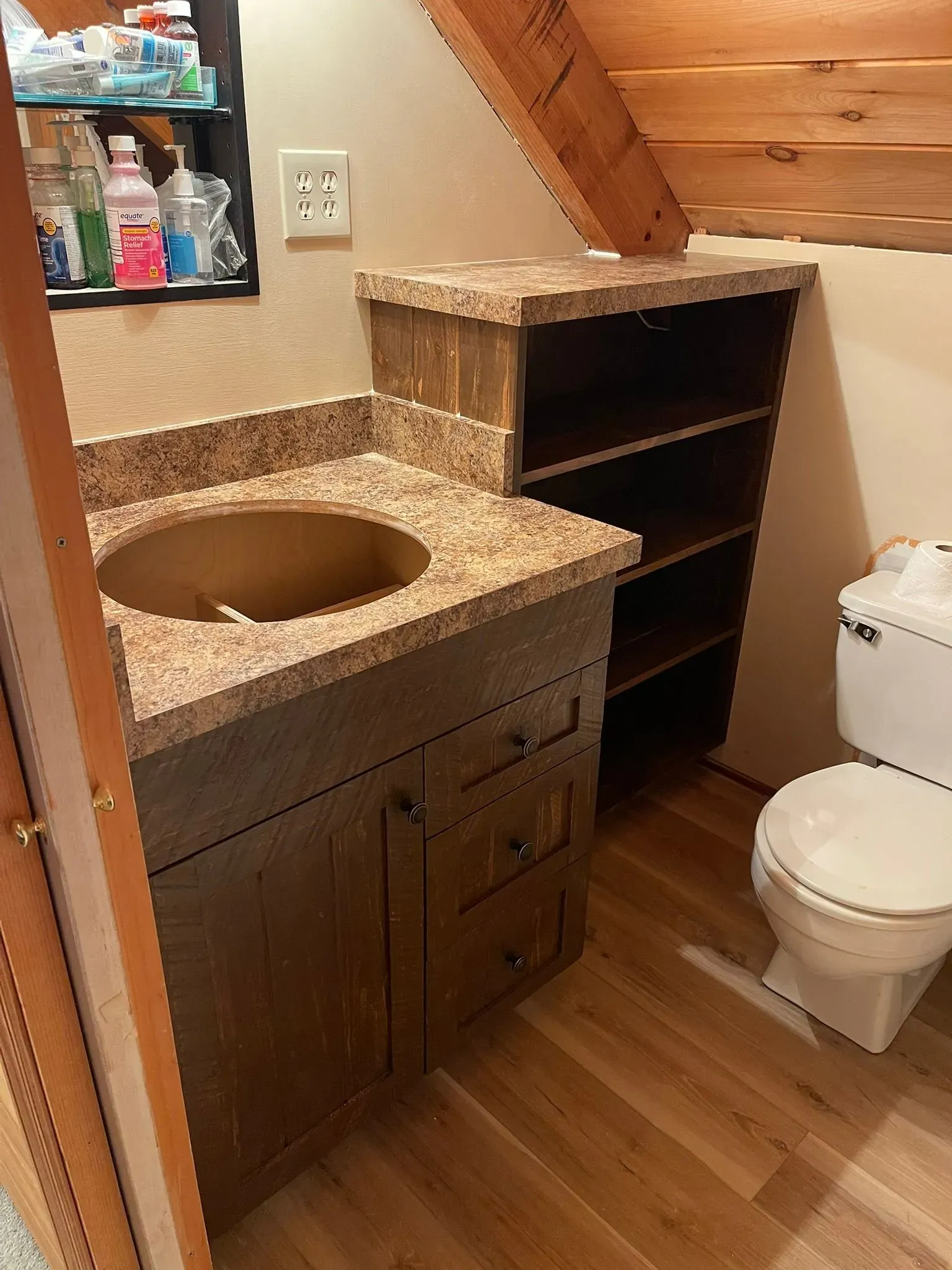 Small bathroom with granite vanity, open shelving, toilet, and slanted wood beam under stairs.