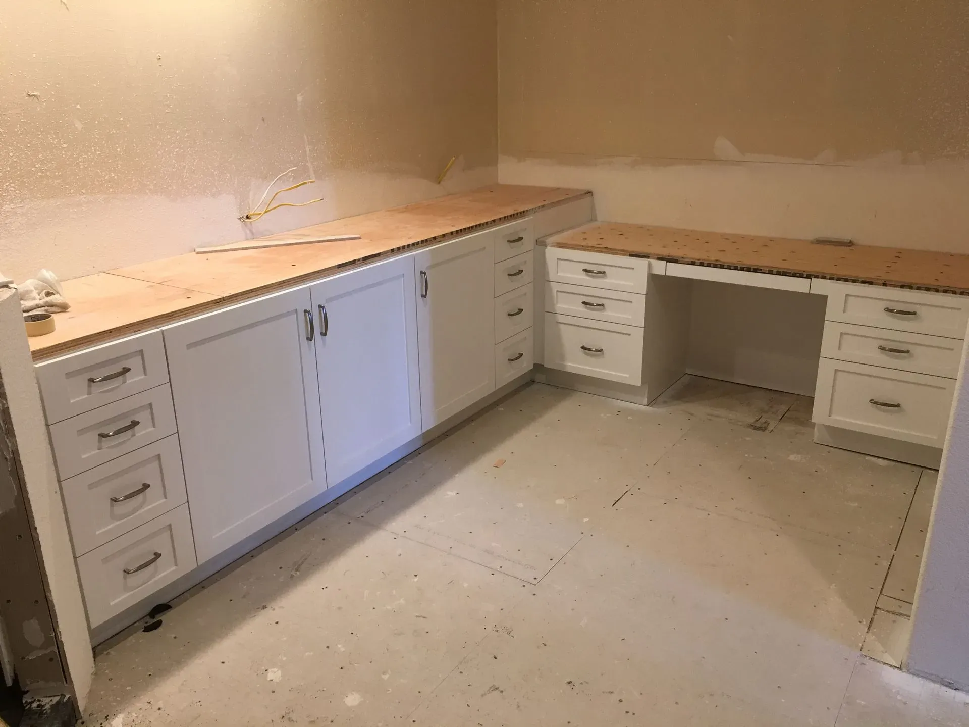 Unfinished kitchen with white cabinets and wooden countertops along two walls, beige walls, and exposed floor debris