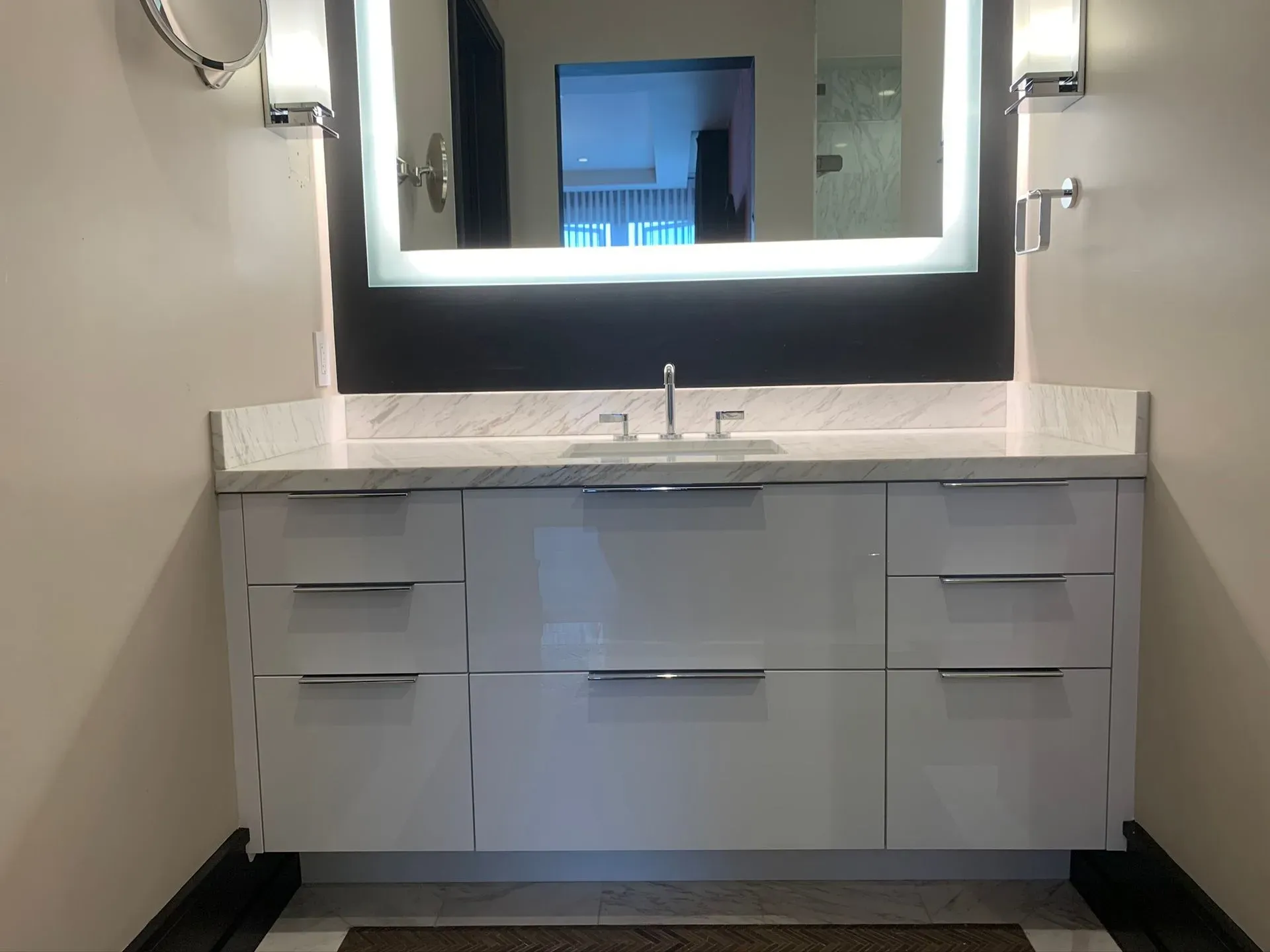 Modern bathroom vanity with white cabinets, marble countertop, and illuminated mirror above the sink.