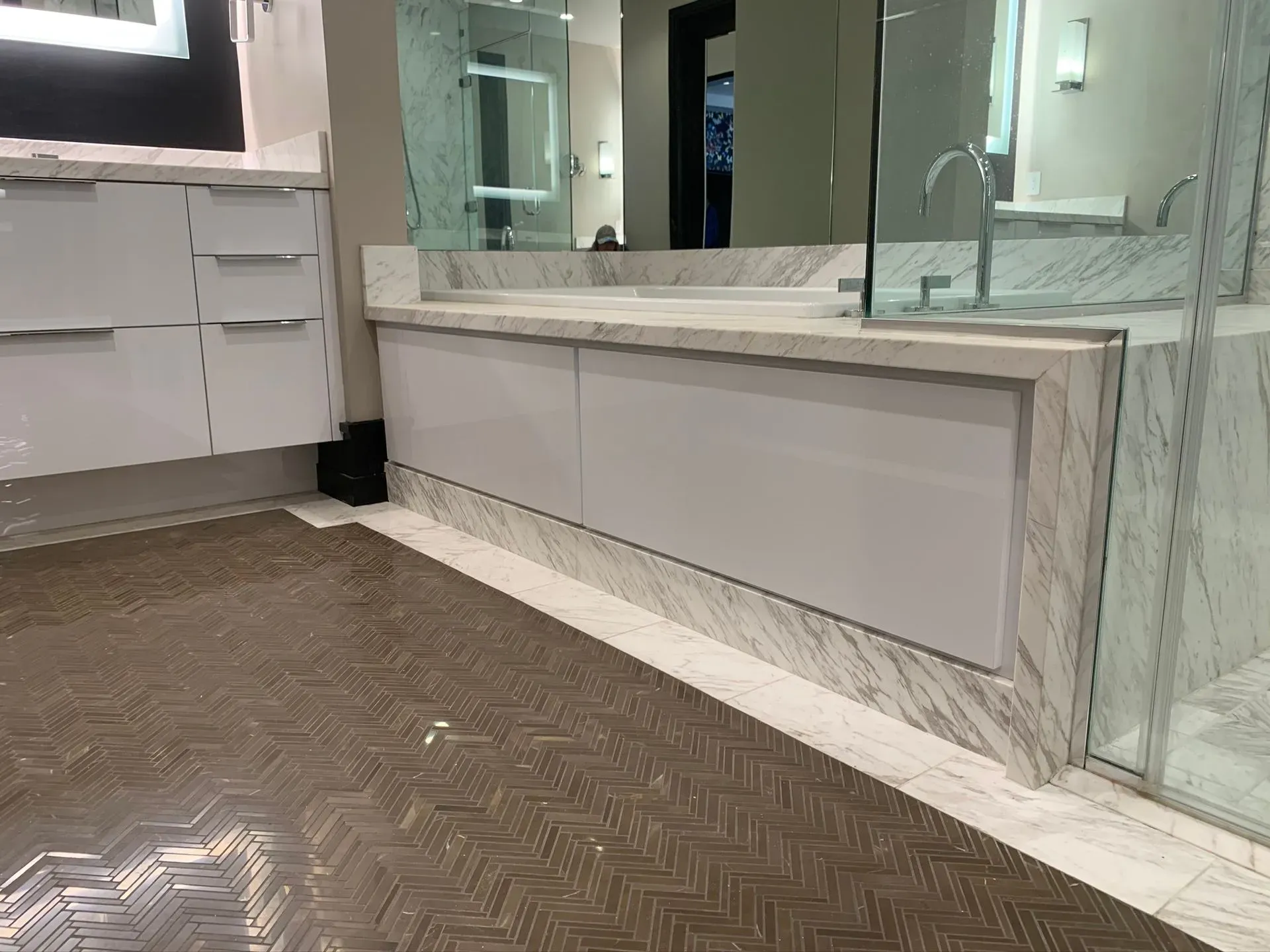 Bathroom vanity with mirrored wall, white marble surfaces, and herringbone tile floor