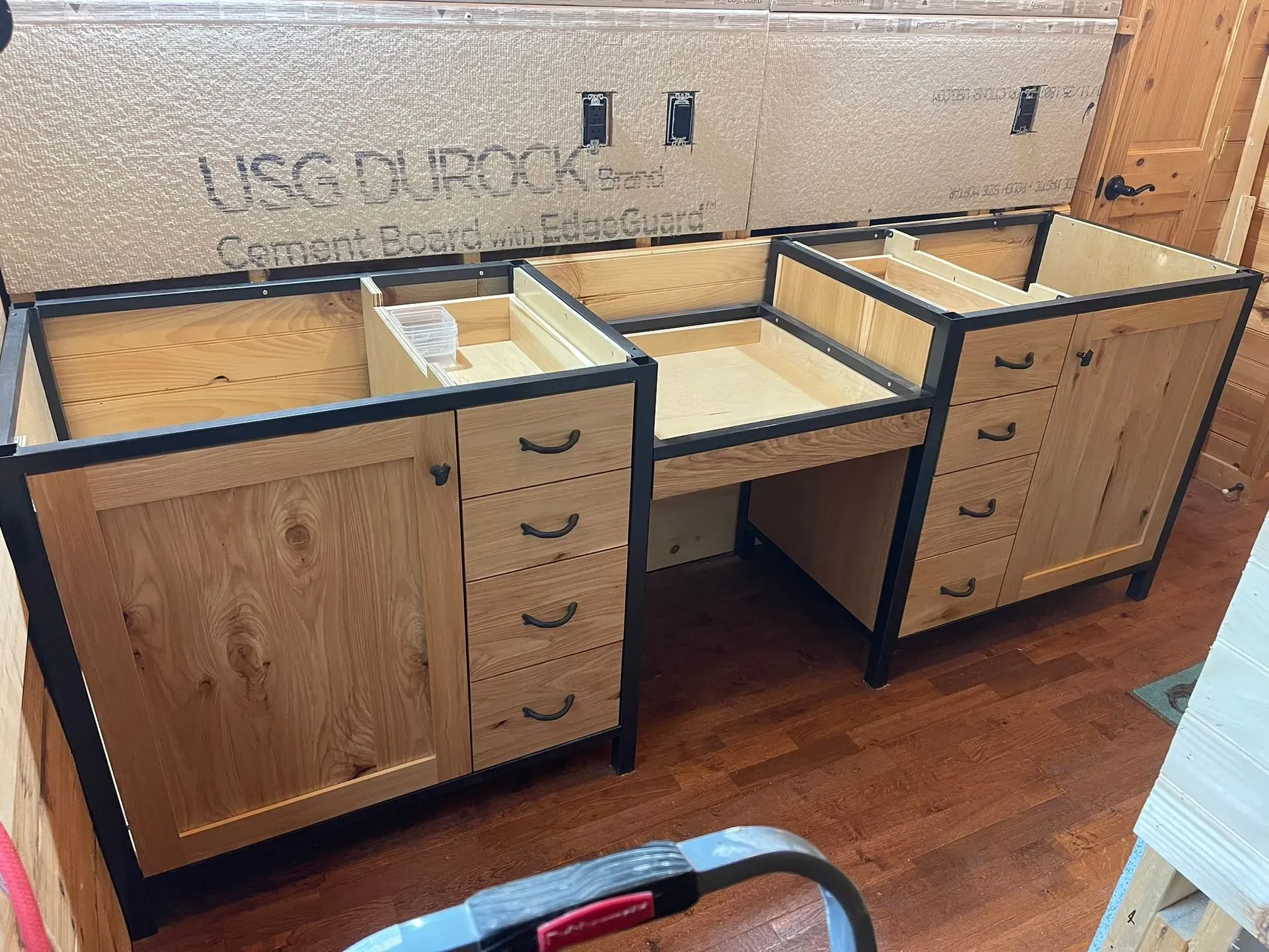 Wooden kitchen cabinets and drawer units laid out in a room, with a wood floor and wall opening behind them.
