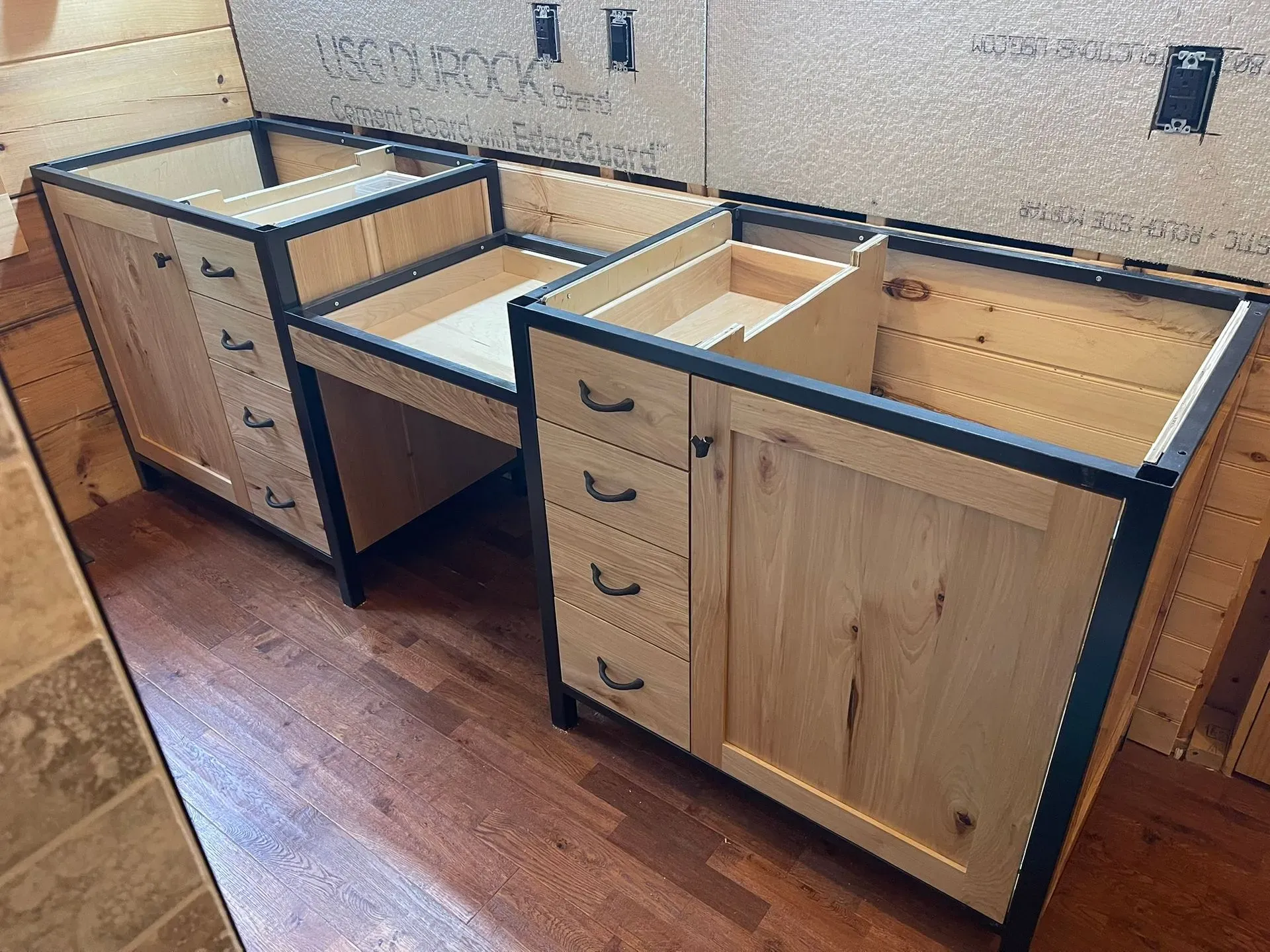 Two unfinished wood kitchen cabinets with dark countertops installed along a wall in a tiled room.