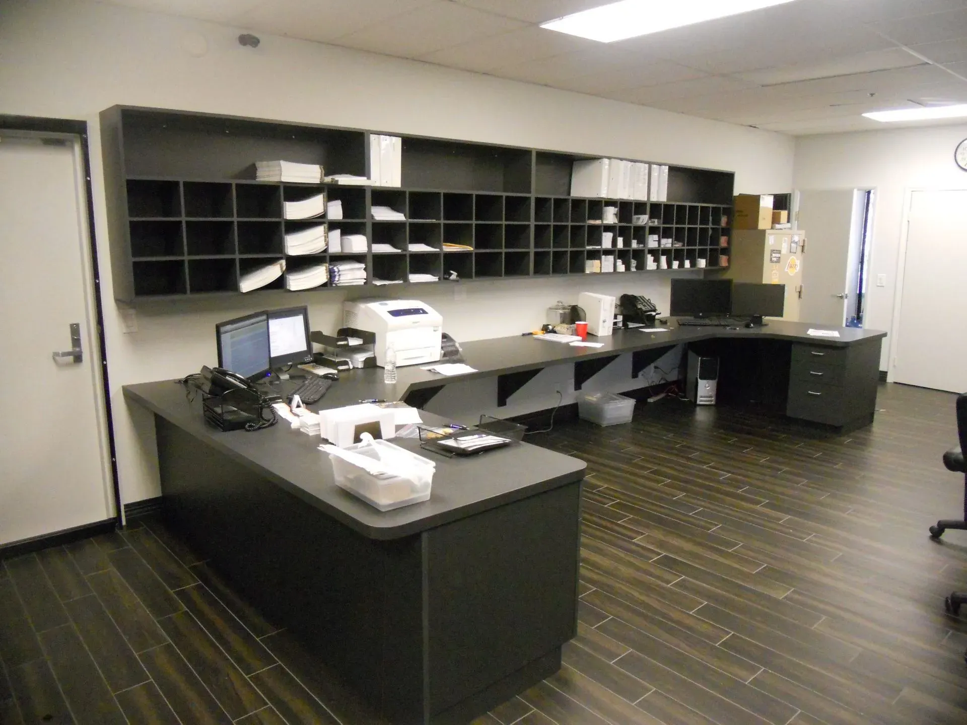 Empty office with desks, cubbies, computers, and a printer under fluorescent lights.