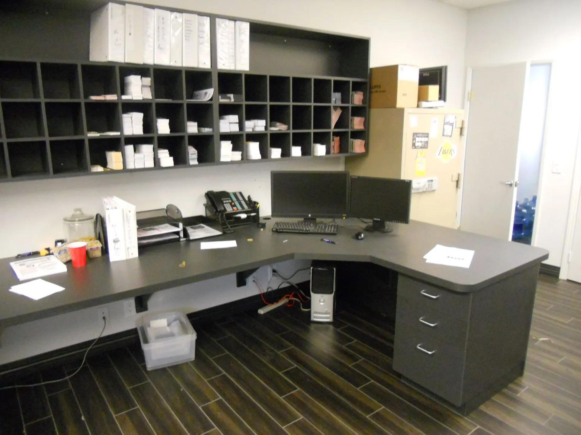 Office workspace with black L-shaped desk, computer monitors, cubbies, and filing cabinets