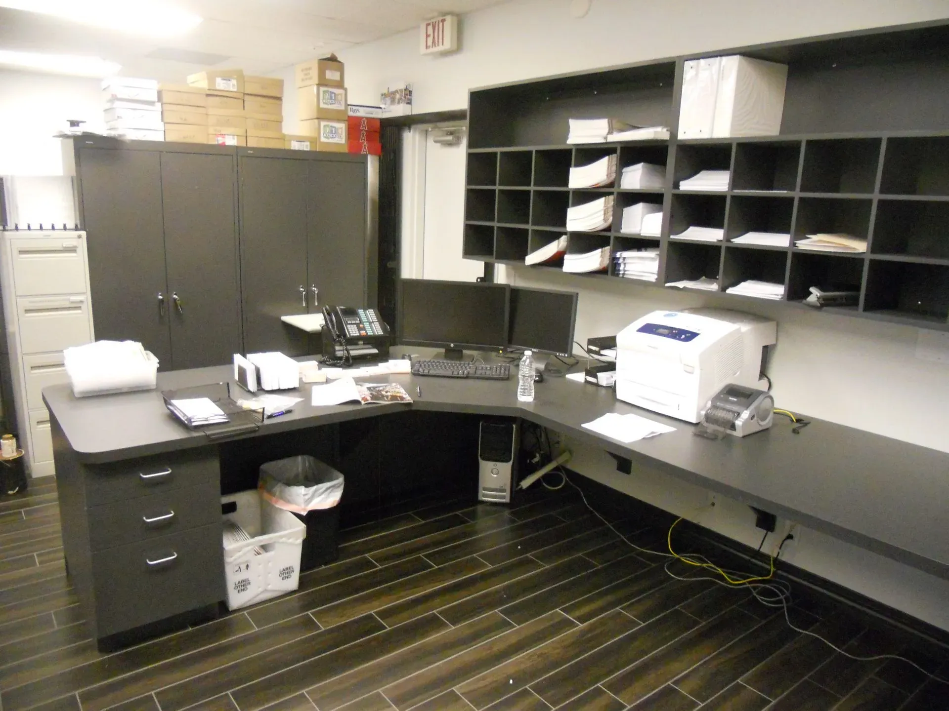 Office with dark cabinets, desk, printer, mail cubbies, and scattered papers on a striped floor.