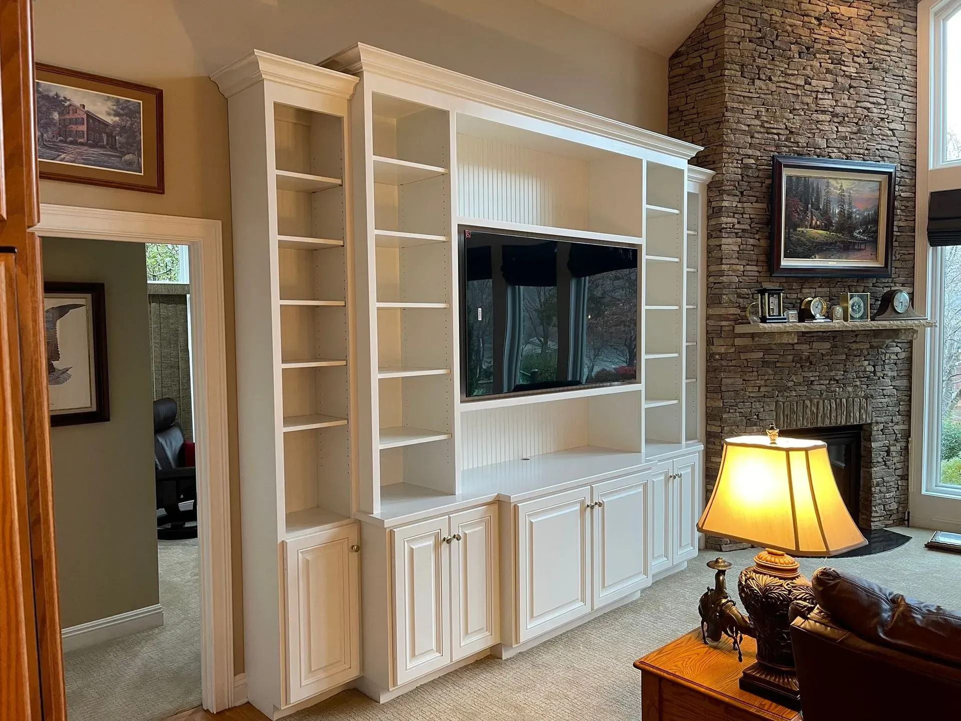 Living room with built-in white cabinets, a wall-mounted TV, stone fireplace, and lit table lamp