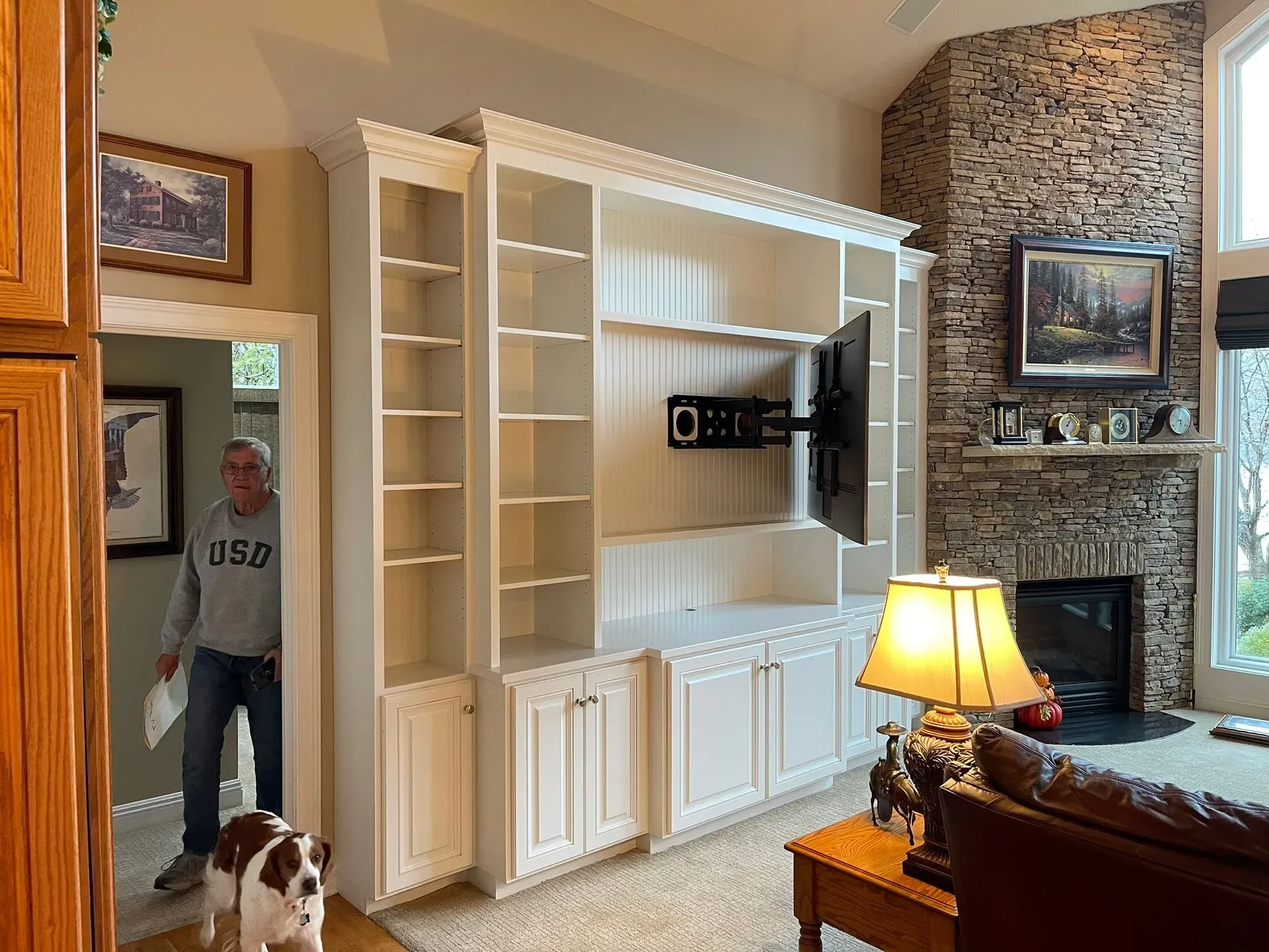Living room with a large white built-in cabinet, stone fireplace, lamp, and a person standing by the doorway with a dog.