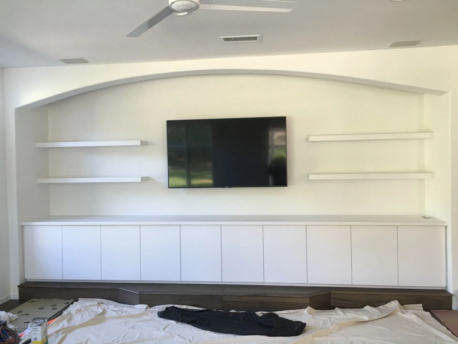 Modern living room wall with mounted TV, floating shelves, and long white cabinets beneath an arched accent wall