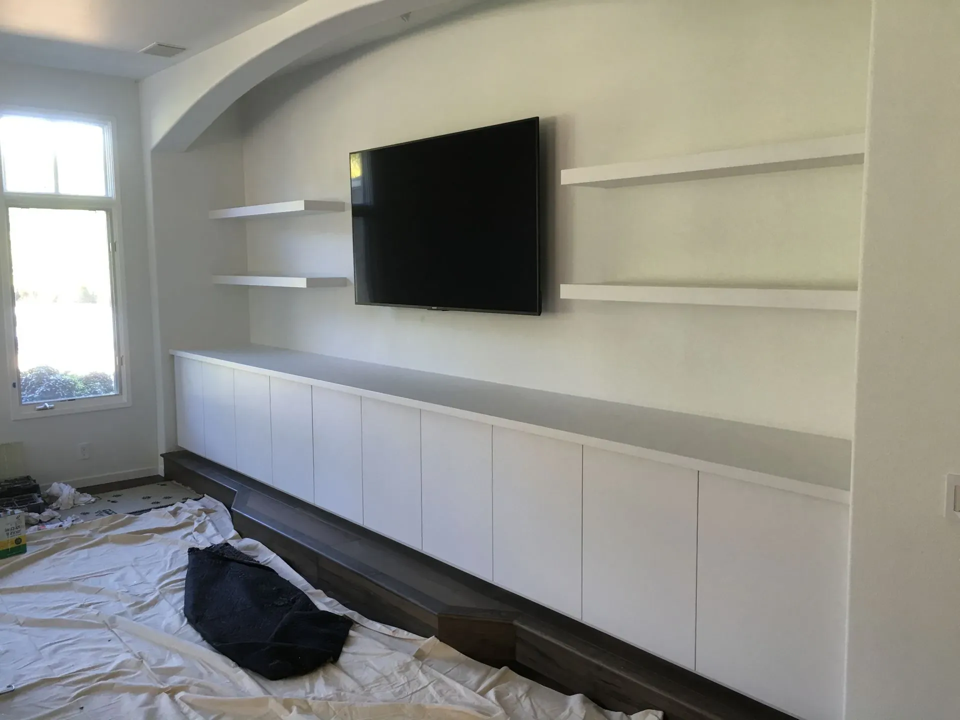 Living room with wall-mounted TV, white built-in cabinets, floating shelves, and drop cloths on the floor