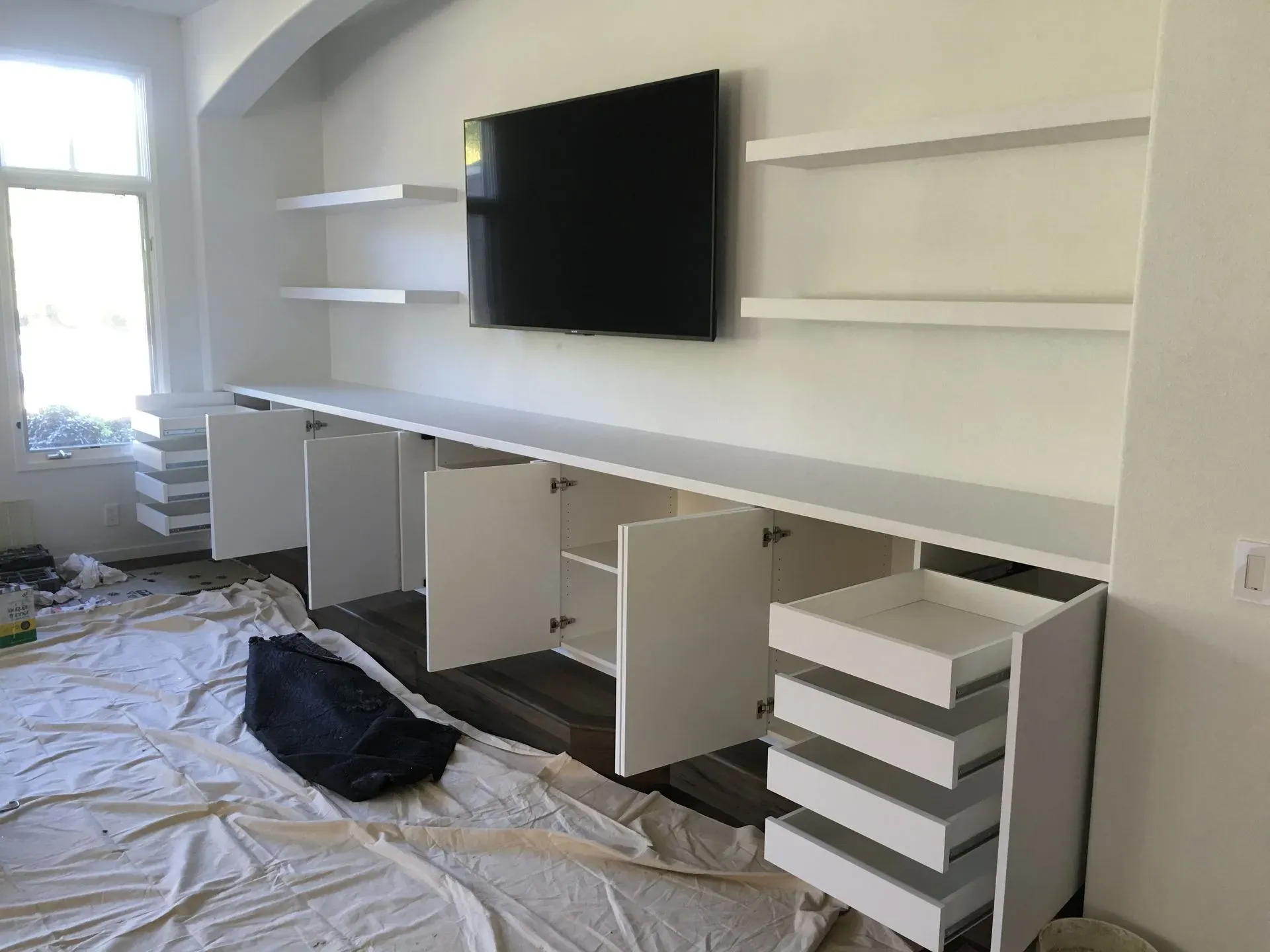 Modern white built-in desk wall with cabinets, shelves, and a mounted TV in a room under renovation.