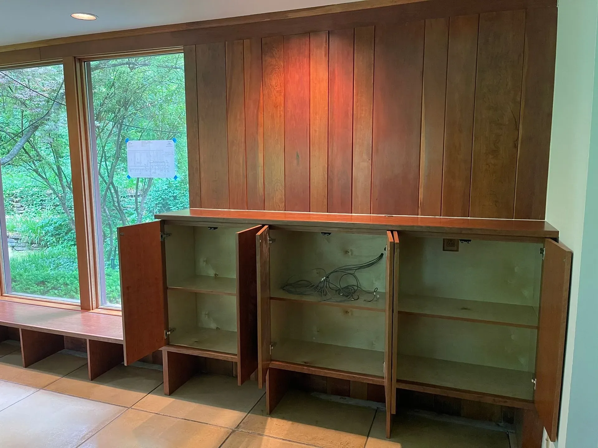 Wood-paneled room with three open cabinets beneath large windows overlooking greenery