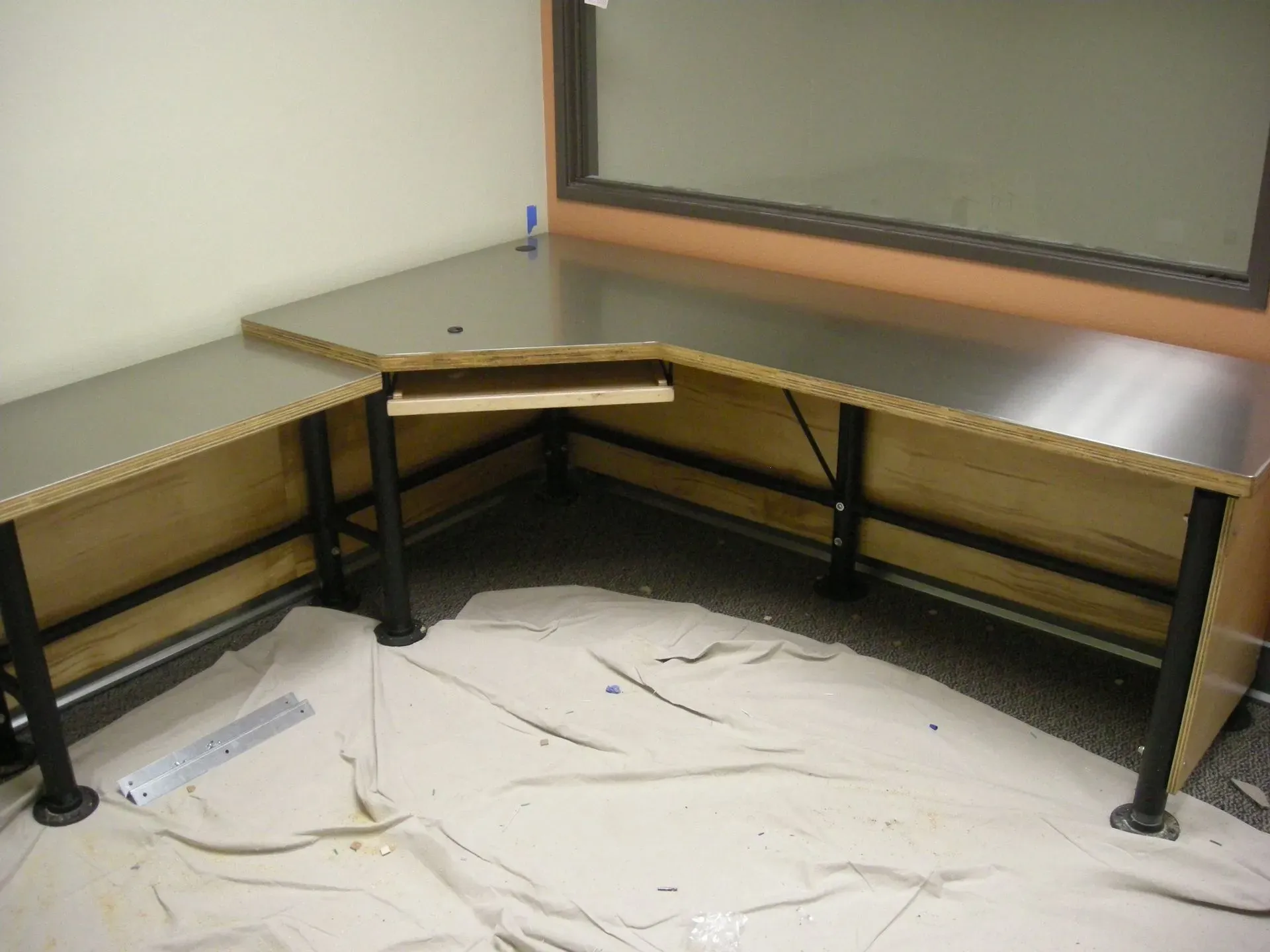 Corner L-shaped desk setup with light wood surfaces, black legs, and a white floor covering in a bare room