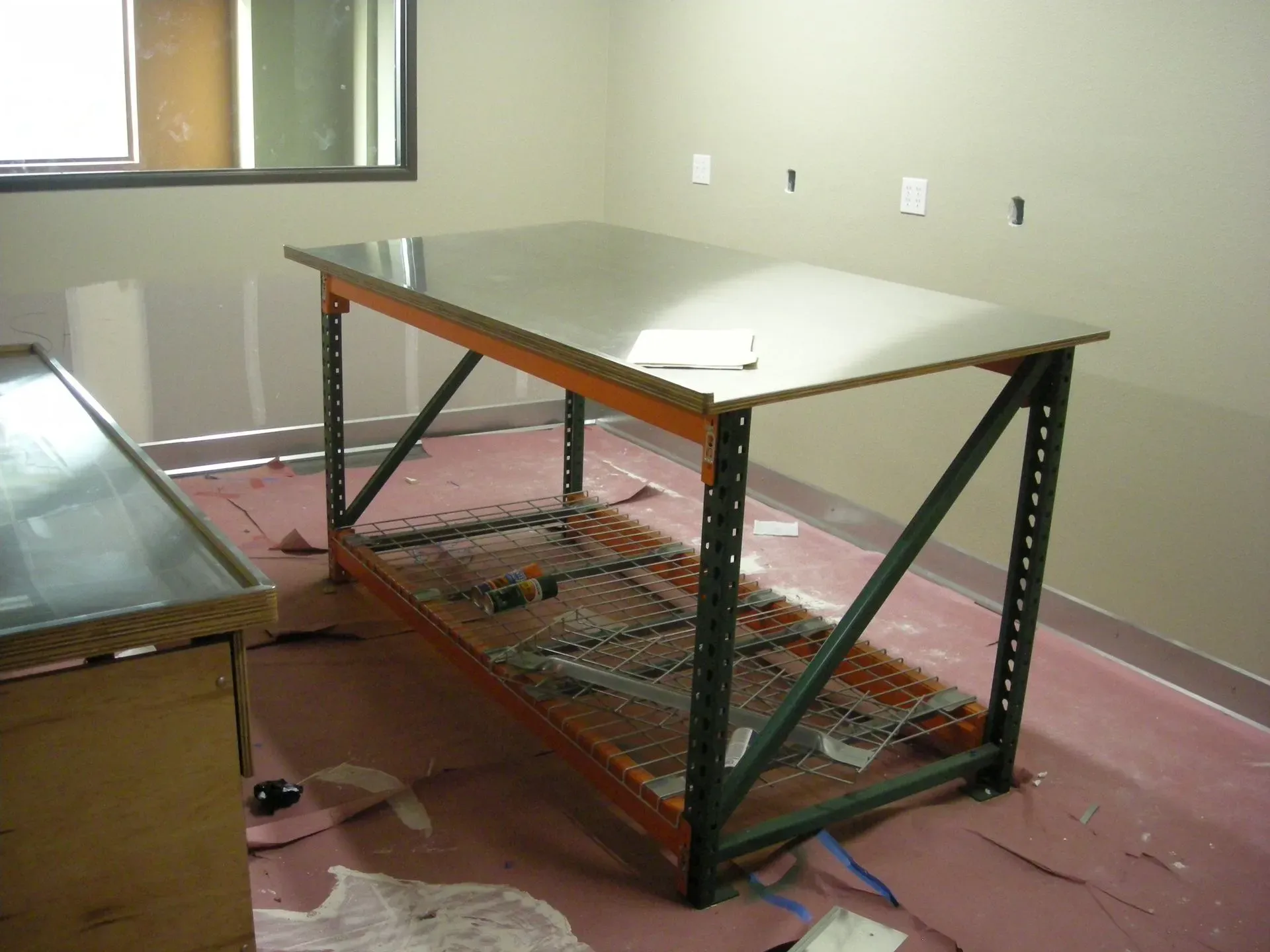 Empty metal workbench in a bare room with wires on the floor and a glass table at left
