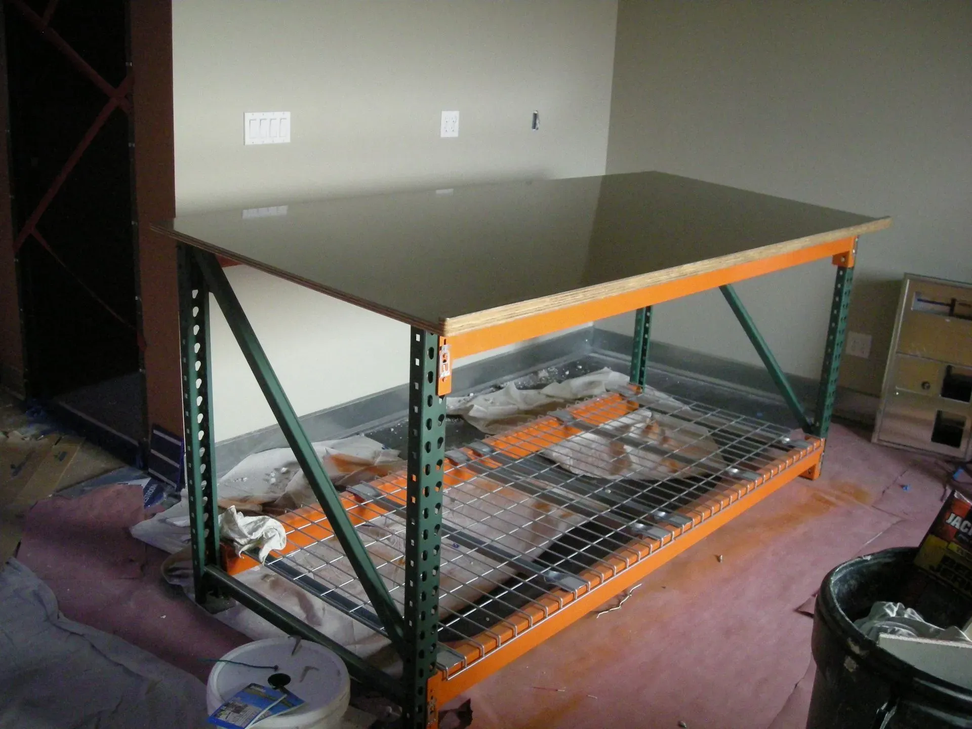 Green metal workbench with a wooden top in a storage area, surrounded by shelves and supplies.