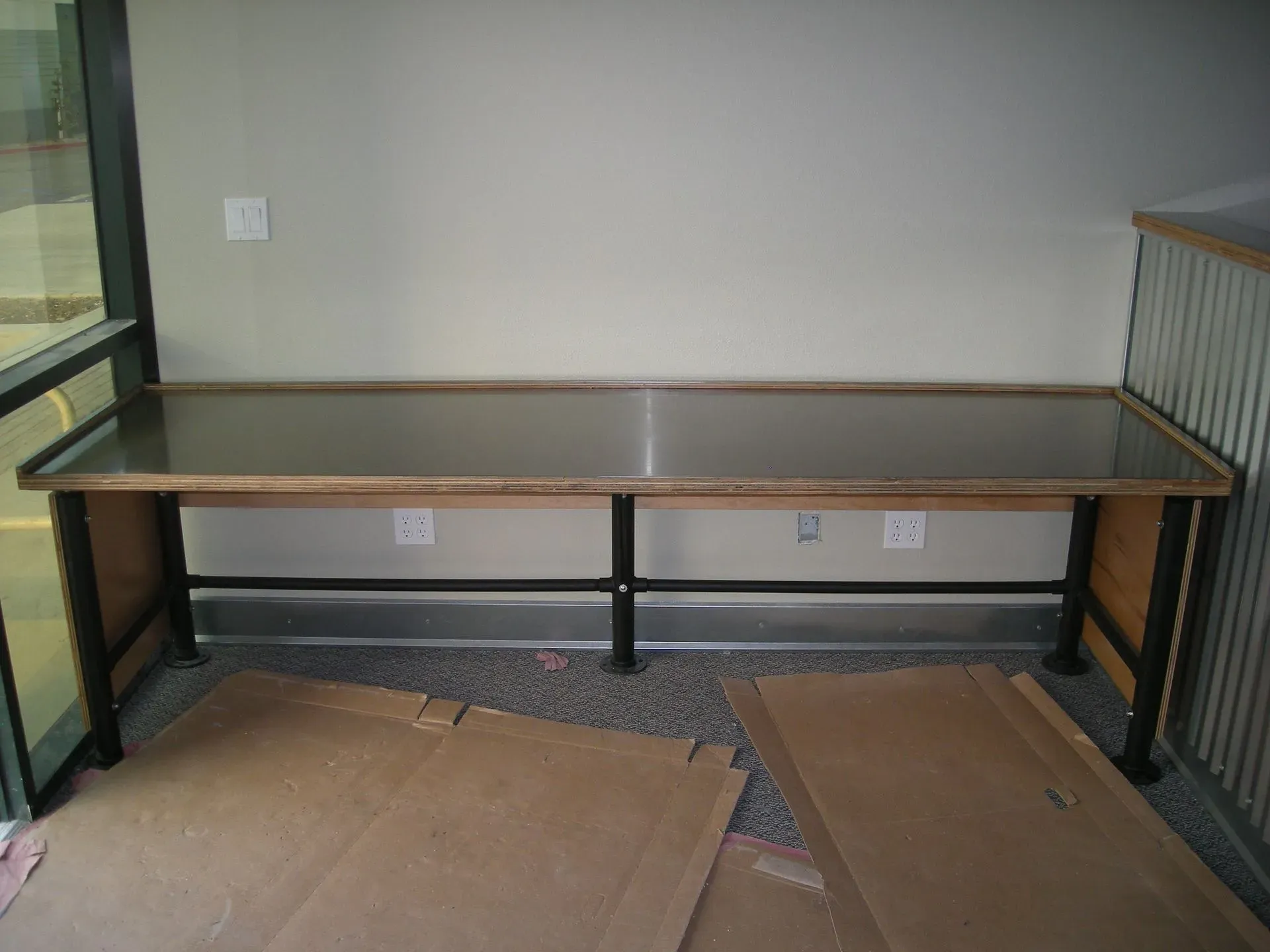 Long wooden desk against a wall in an office area, with cardboard pieces on the floor in front.