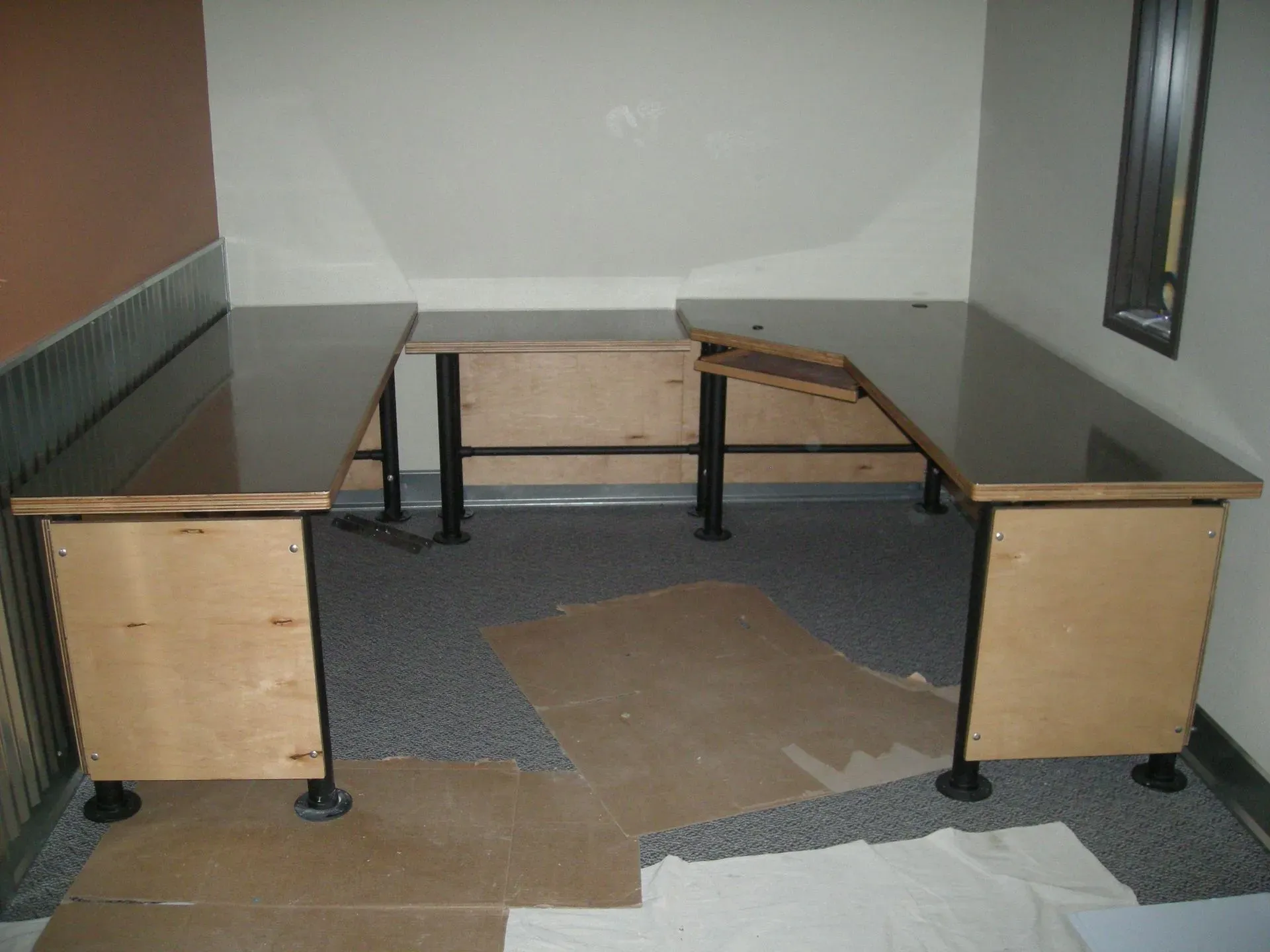 L-shaped desk in an empty office corner with gray and wood finishes and a damaged floor mat.