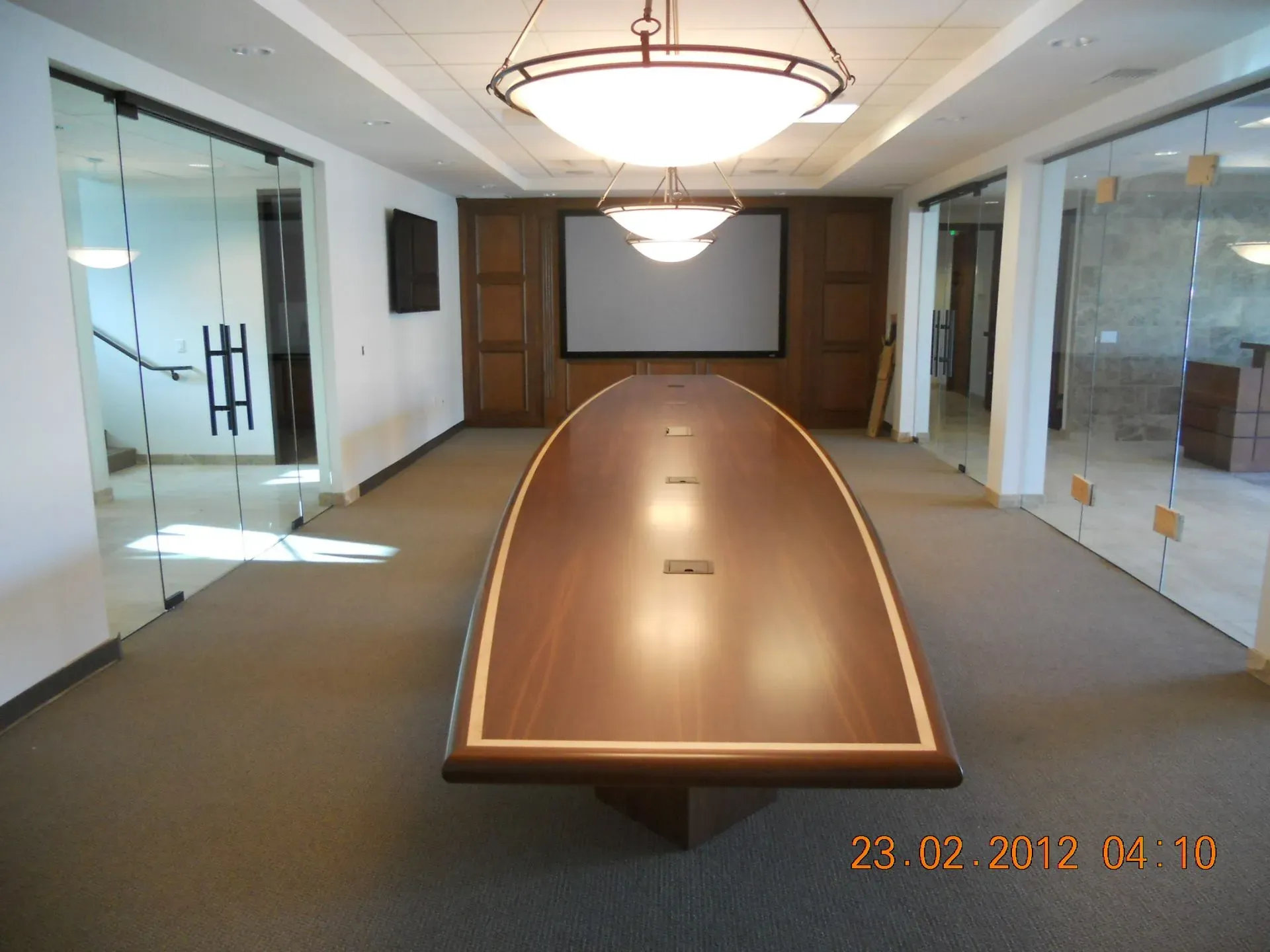 Conference room with a long wooden table, glass walls, and a projector screen at the far end.