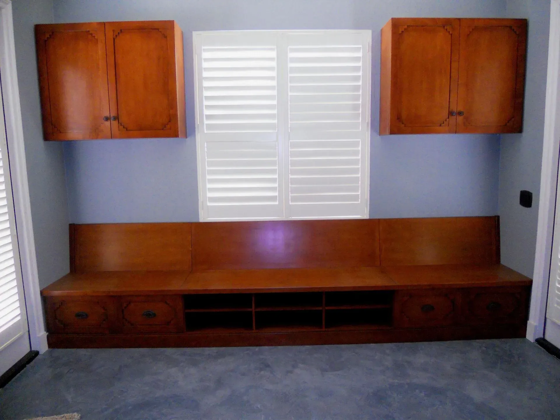 Built-in wooden entertainment center with cabinets and drawers against a wall with window shutters