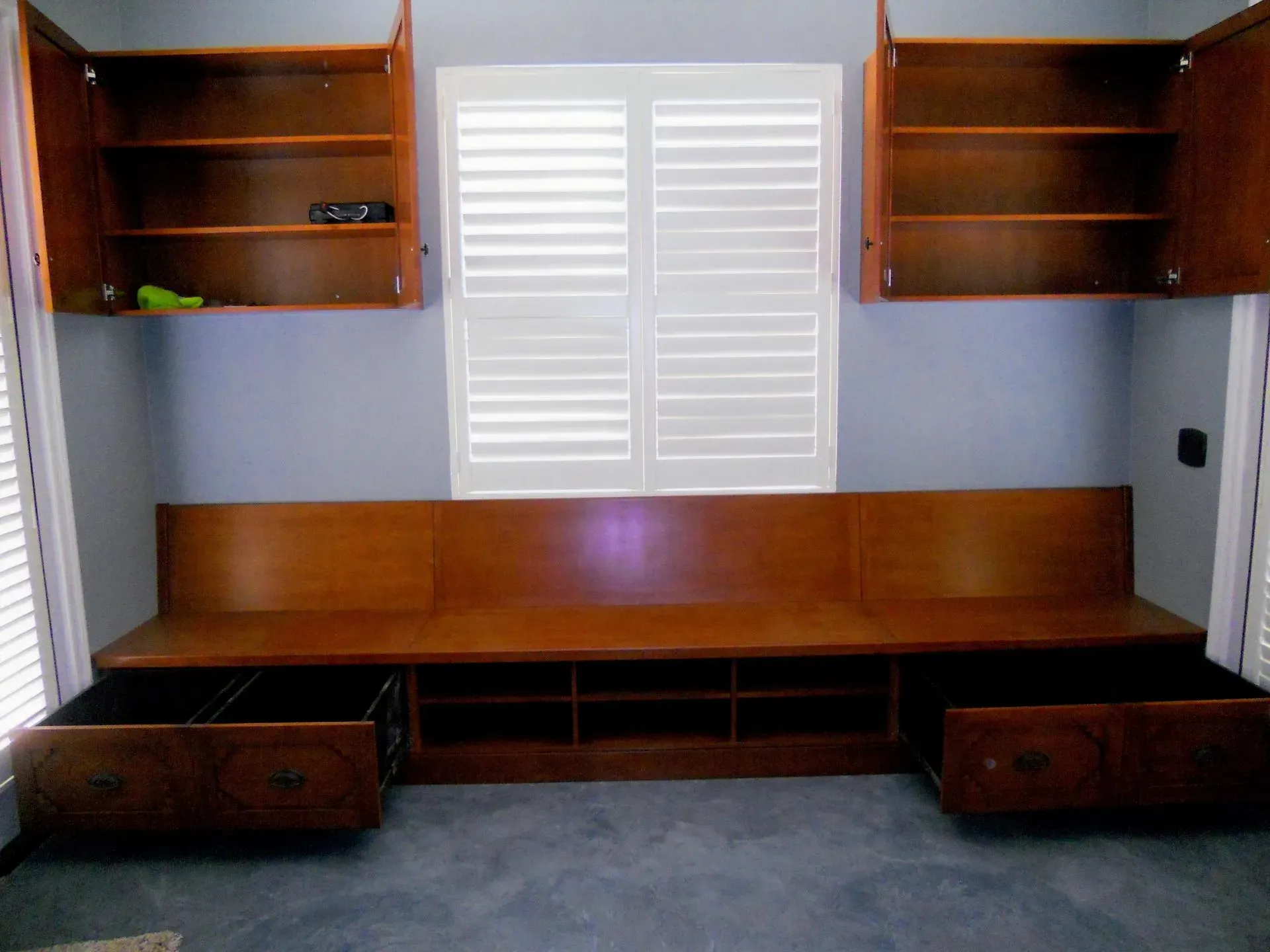 Built-in wooden desk with shelves and drawers beneath a white shuttered window