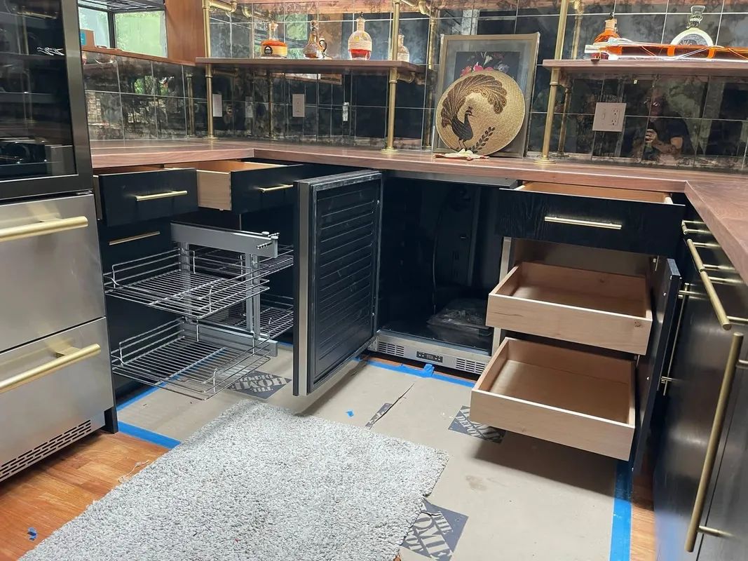 Open dishwasher with racks and trays under a kitchen counter, flanked by a fridge and drawers.