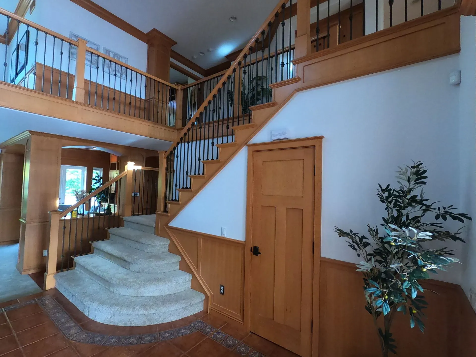Interior foyer with curved staircase, wooden railings, and a wooden door beneath a lofted balcony.