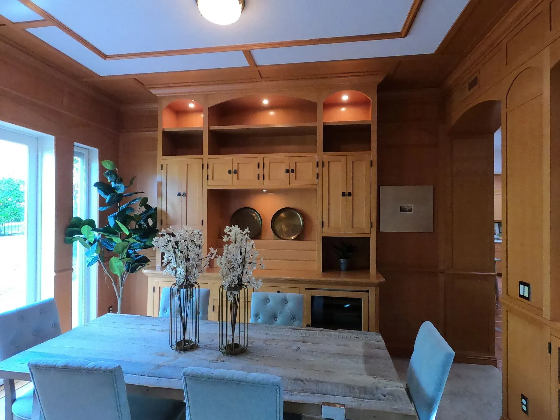 Warm wood-paneled dining room with a table, blue chairs, and built-in shelving by a window