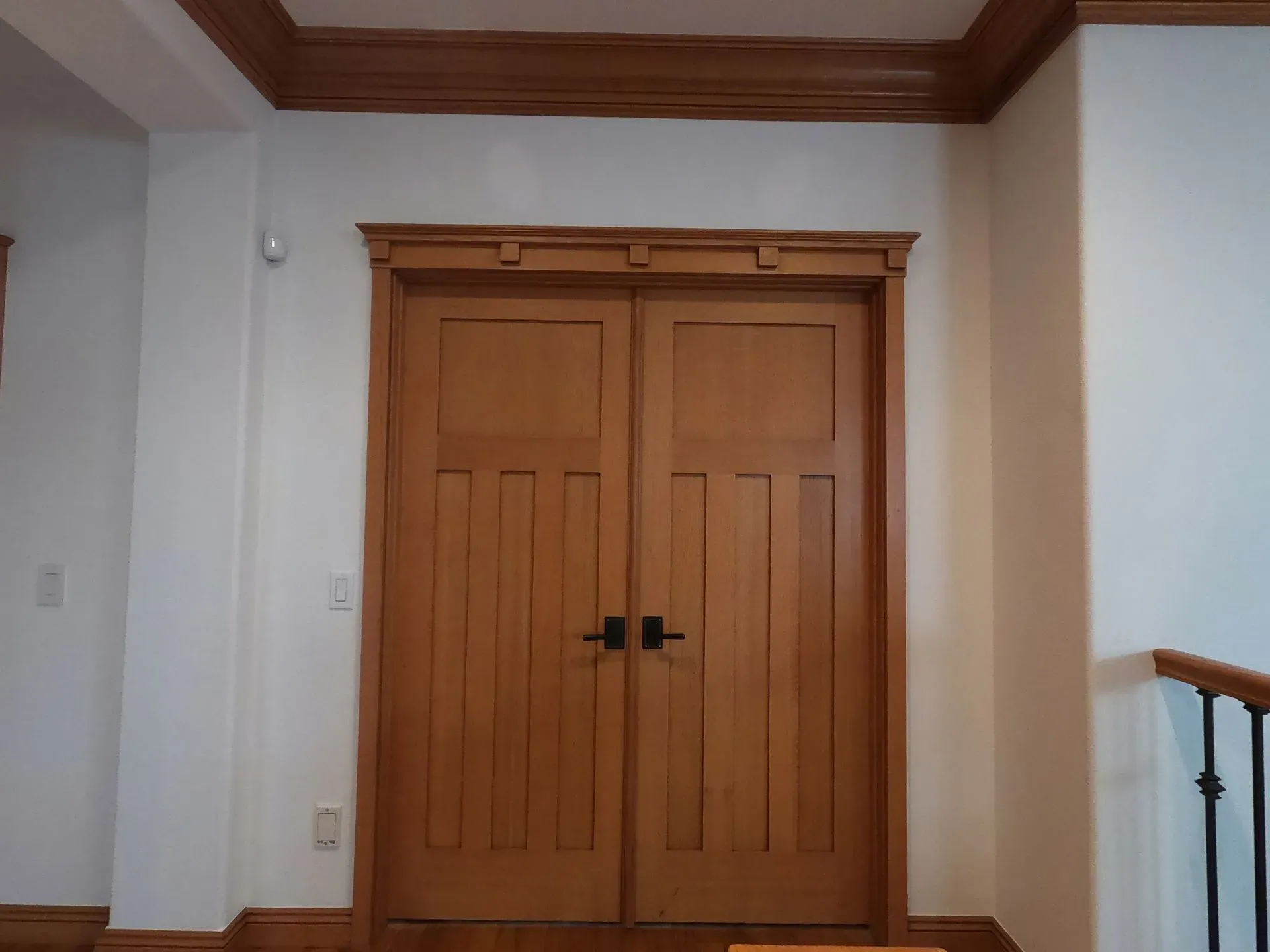 Wooden double doors with black handles in a white hallway with crown molding and a stair railing.