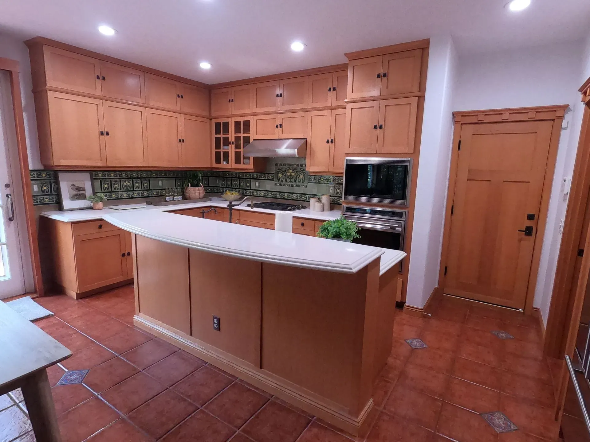 Spacious kitchen with wooden cabinets, terracotta tile floor, and a white island countertop in warm lighting
