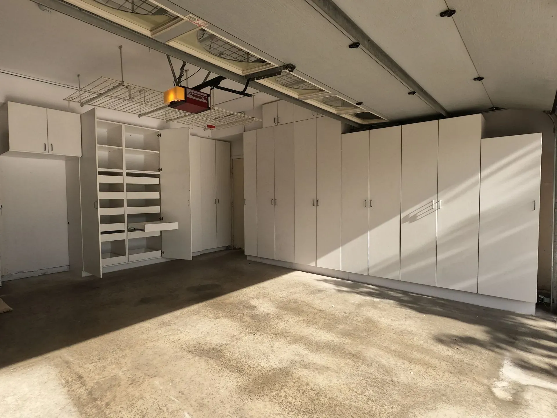 Empty garage interior with white storage cabinets and shelves, sunlight casting shadows on the floor.