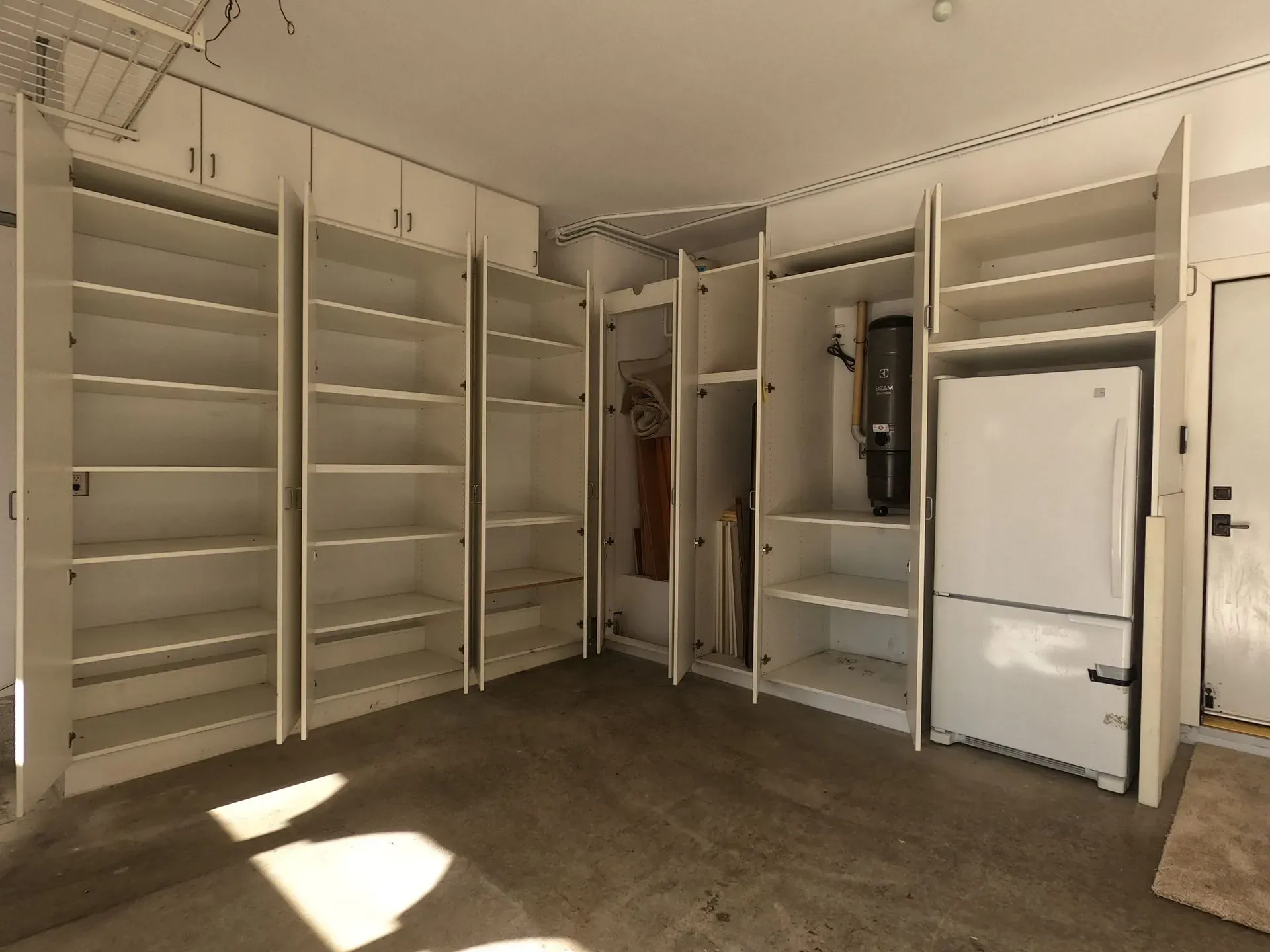 Empty walk-in closet with white built-in shelves and hanging rods, beige carpet, and sunlight on the floor