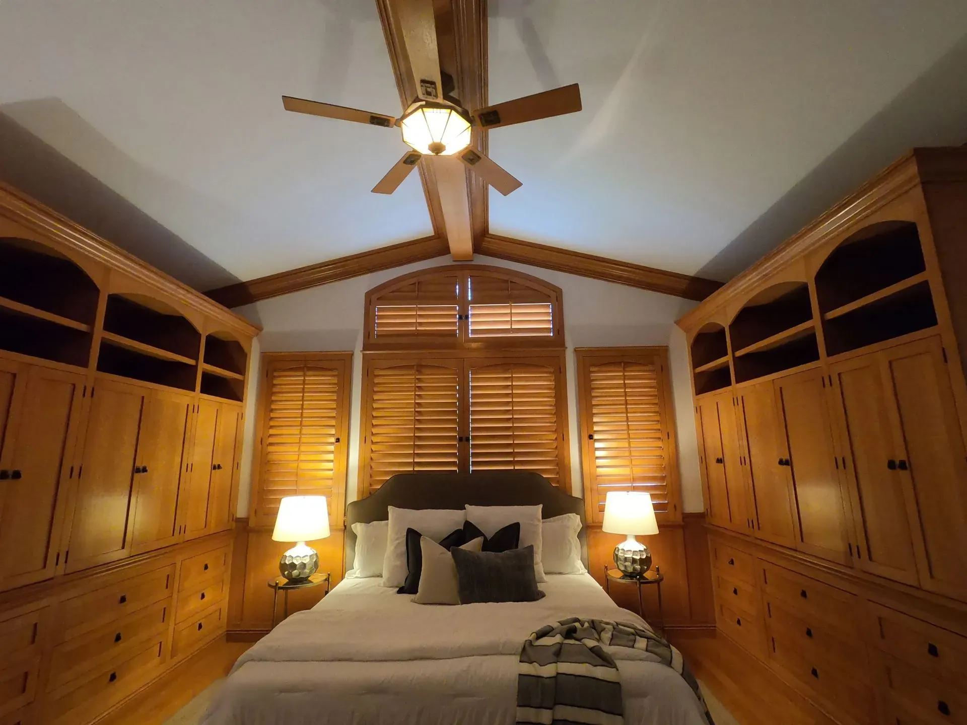 Cozy bedroom with wood-paneled walls, built-in cabinets, a ceiling fan, and a bed centered under shuttered windows.