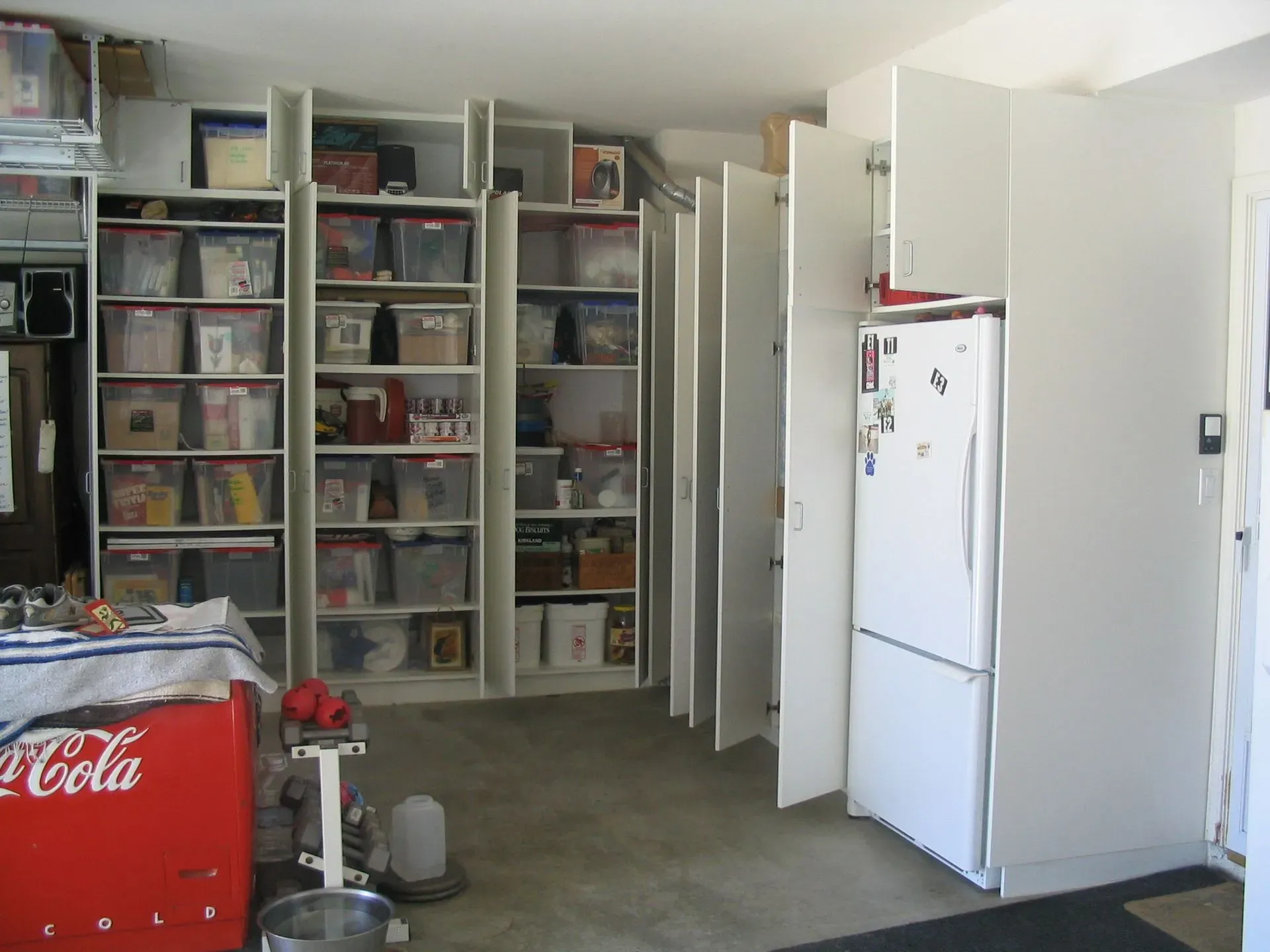 Garage storage room with white cabinets, open shelves of bins, and a white refrigerator