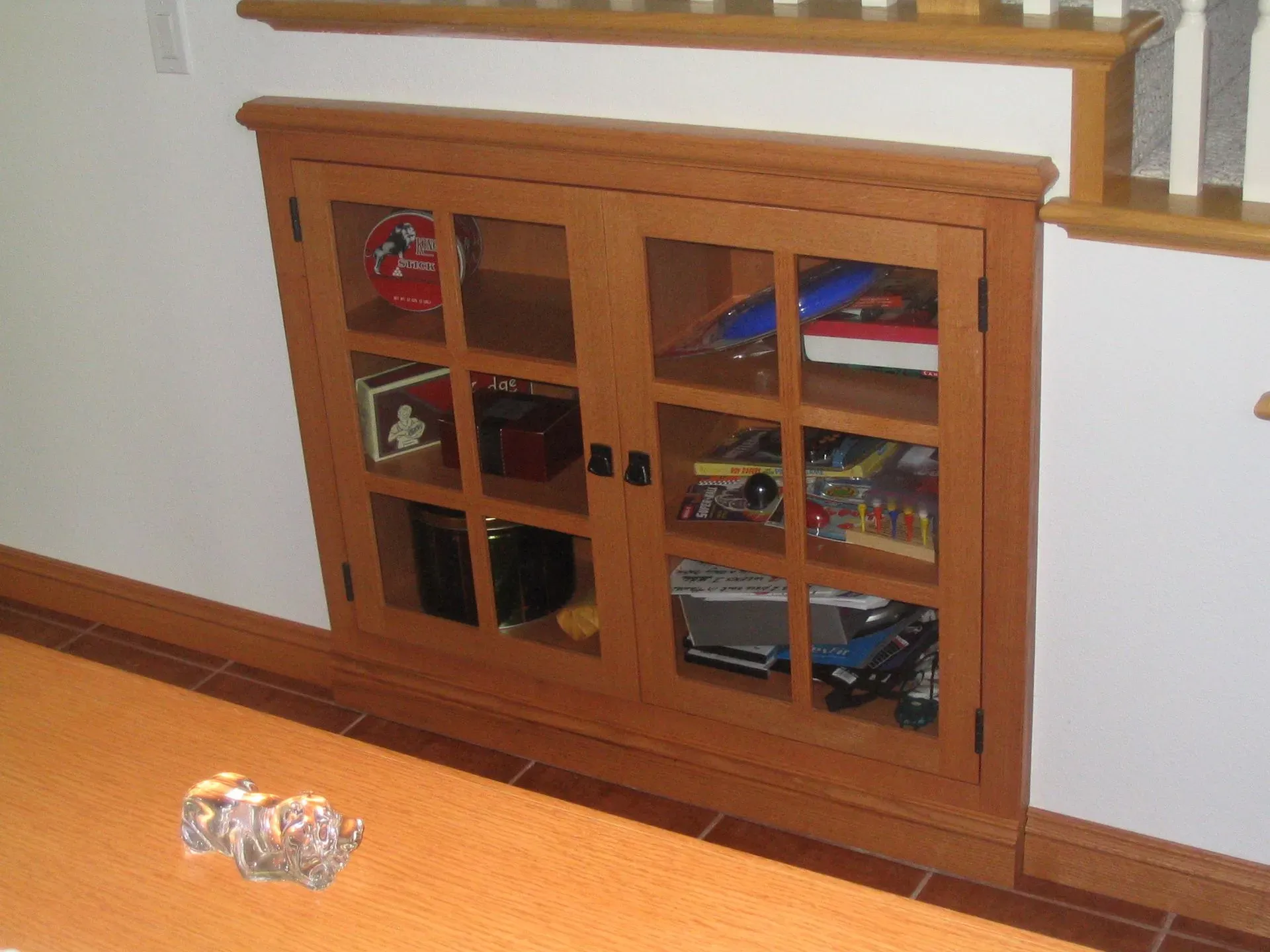 Wooden cabinet with glass doors against a wall, holding books and magazines; a plastic bottle lies on the floor.