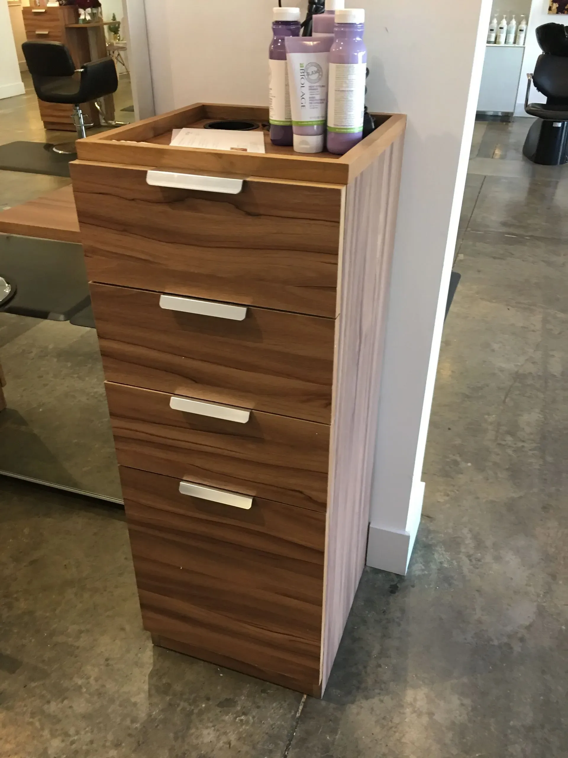 Wooden salon cart with drawers and hair product bottles on top, beside a white wall on a polished floor.