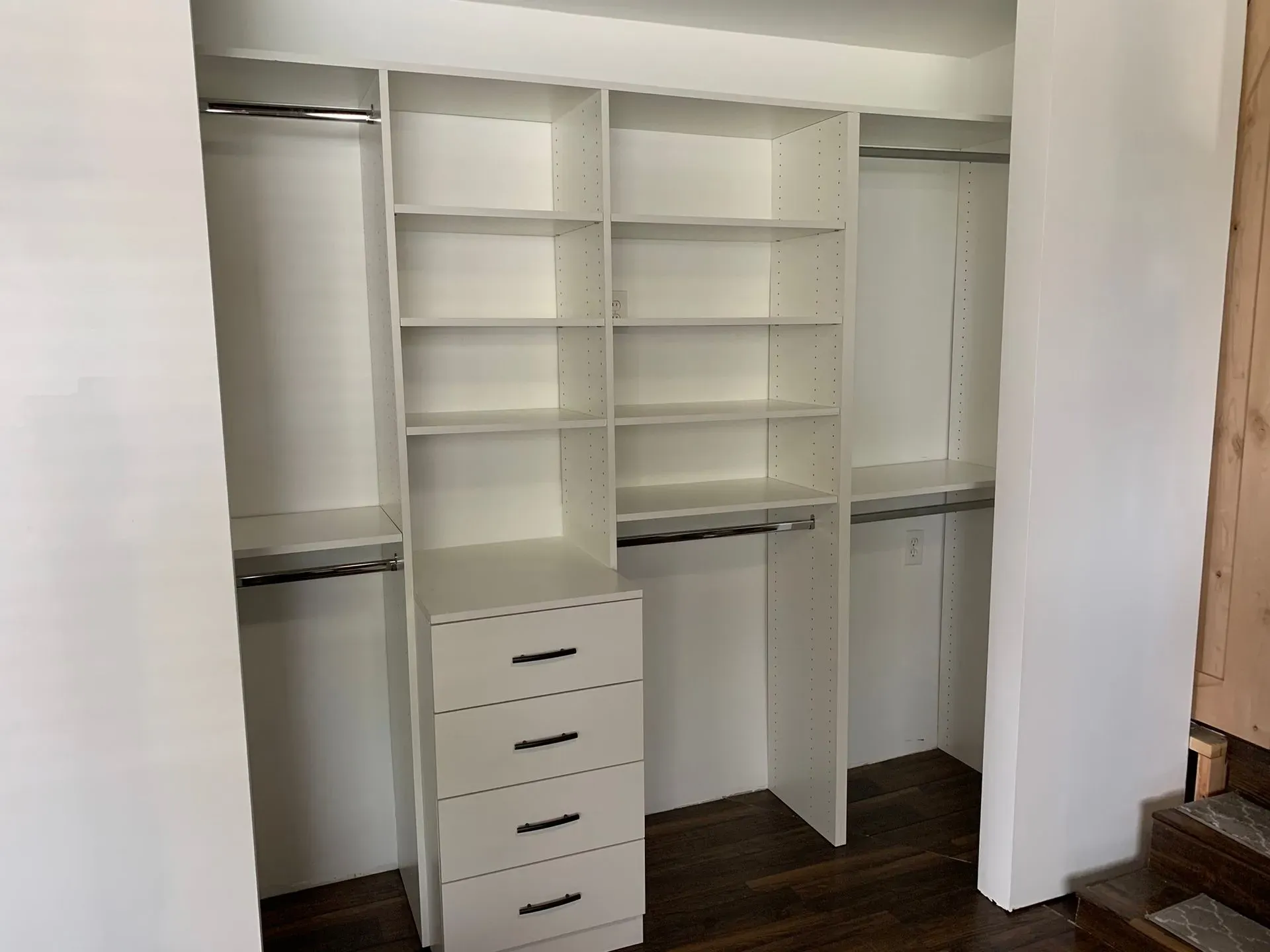 Empty walk-in closet with white built-in shelves, drawers, and hanging rods on dark wood flooring.