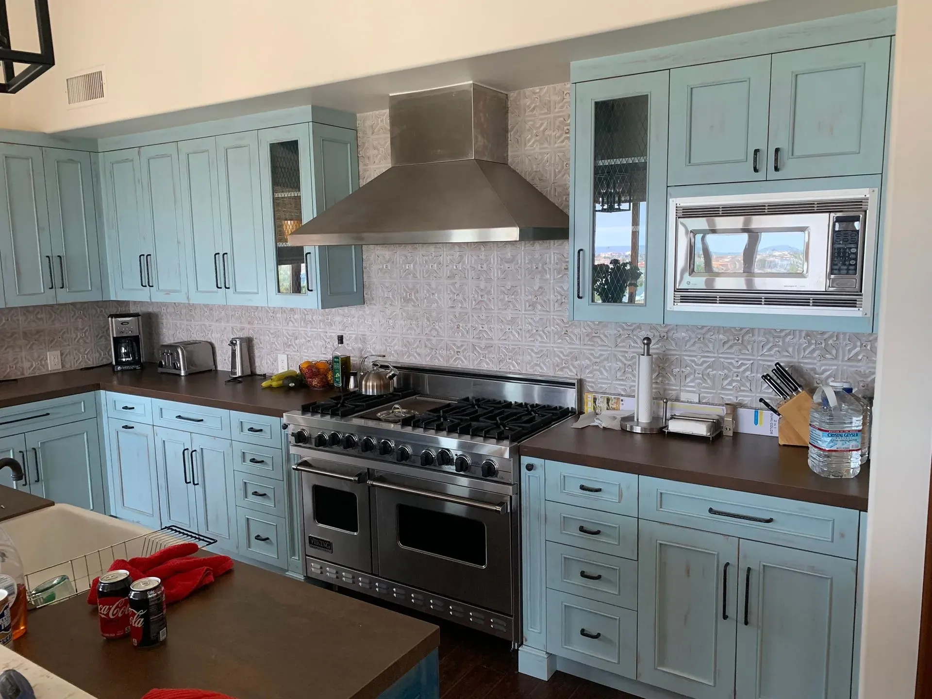 Bright kitchen with pale blue cabinets, stainless steel range hood, and dark countertops by a window.