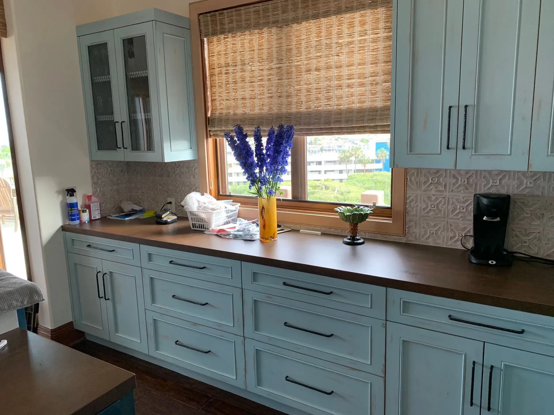 Kitchen with light blue cabinets, dark countertop, window blind, and a vase of purple flowers on the counter