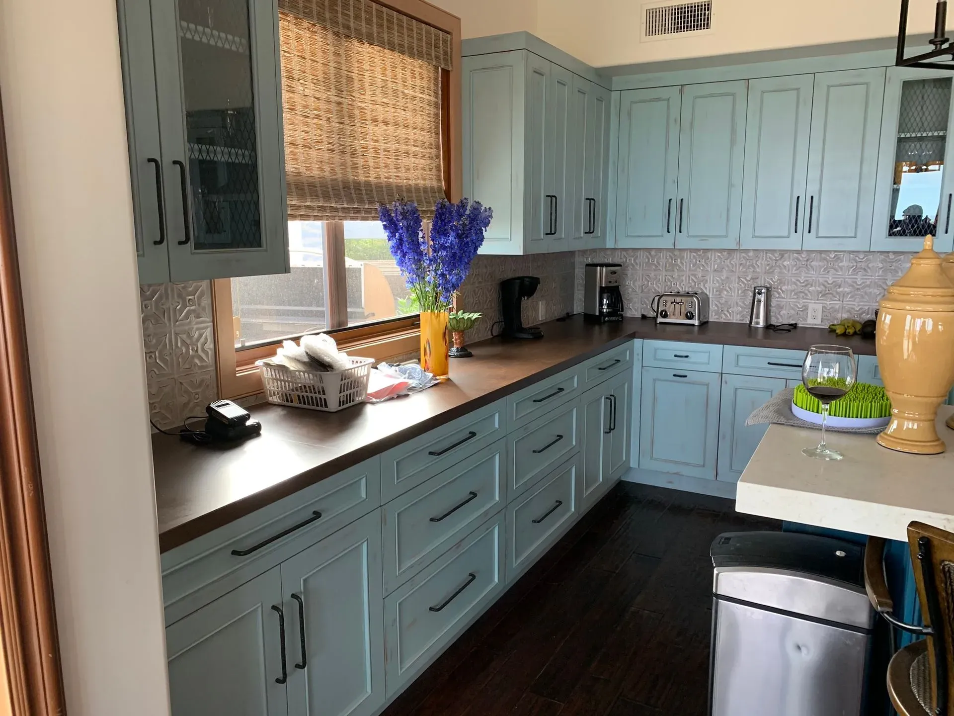 Blue kitchen with black countertops, window blinds, flowers on the counter, and a stainless-steel dishwasher