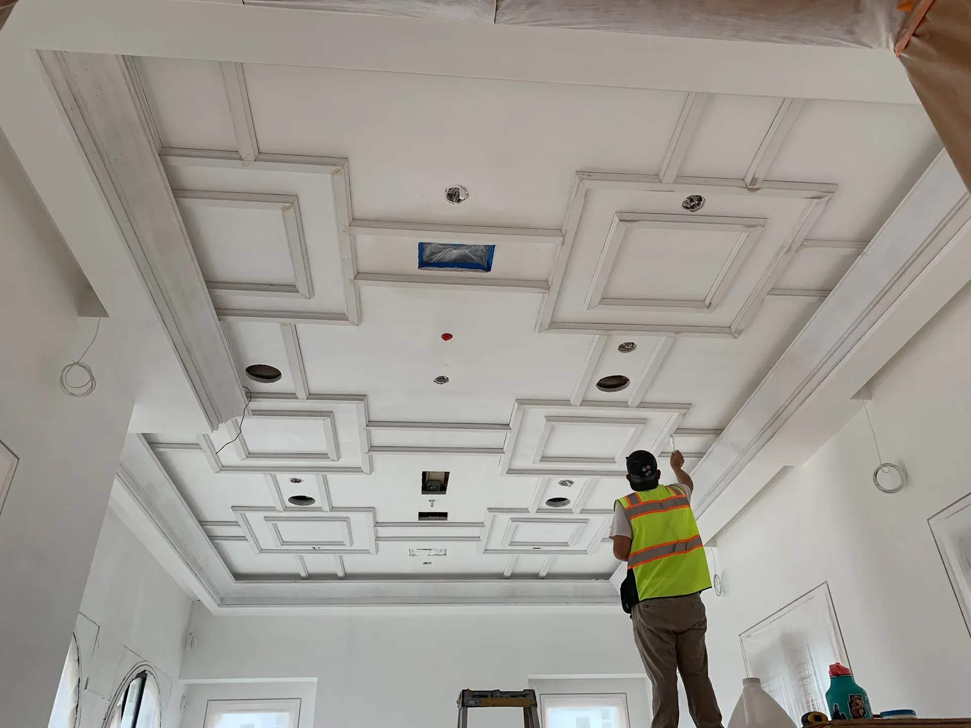 Worker in a yellow safety vest inspecting a decorative ceiling in a white room.