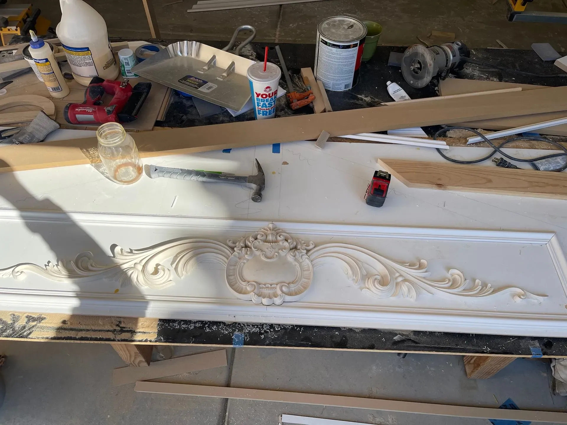 White ornate carved panel lying on a messy workshop floor with tools and paint cans nearby