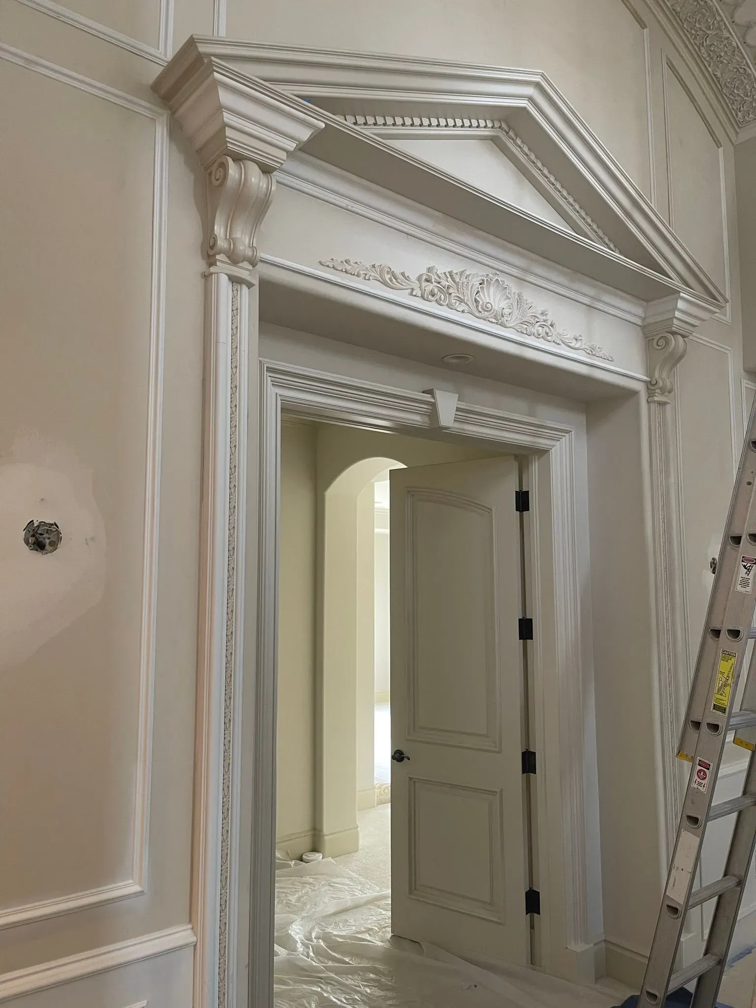 Elegant white doorway with ornate molding, open door, and ladder in a renovated interior room