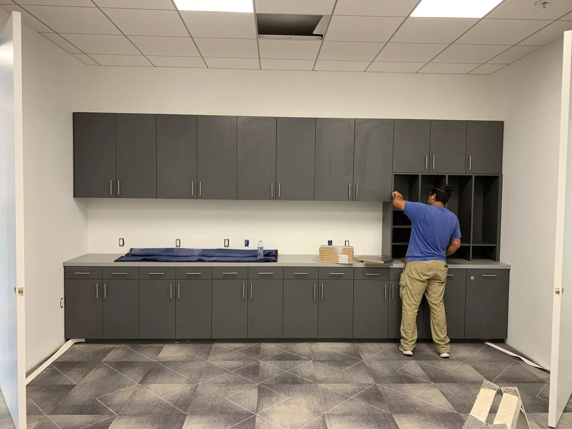 Man in blue shirt installing cabinets in a gray office break room with countertop and sink.