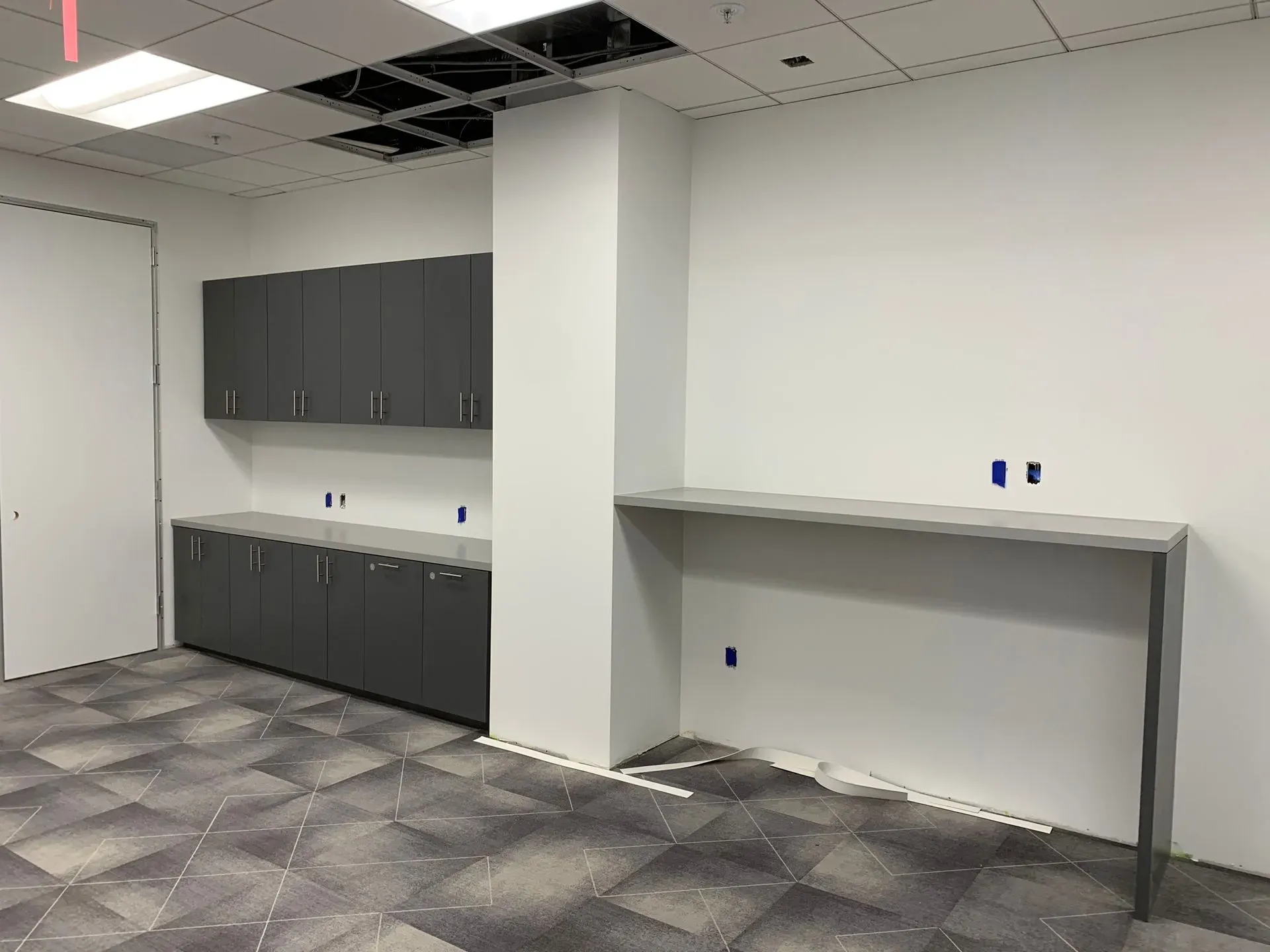 Empty office breakroom with gray cabinets, sink, and a long countertop against white walls.