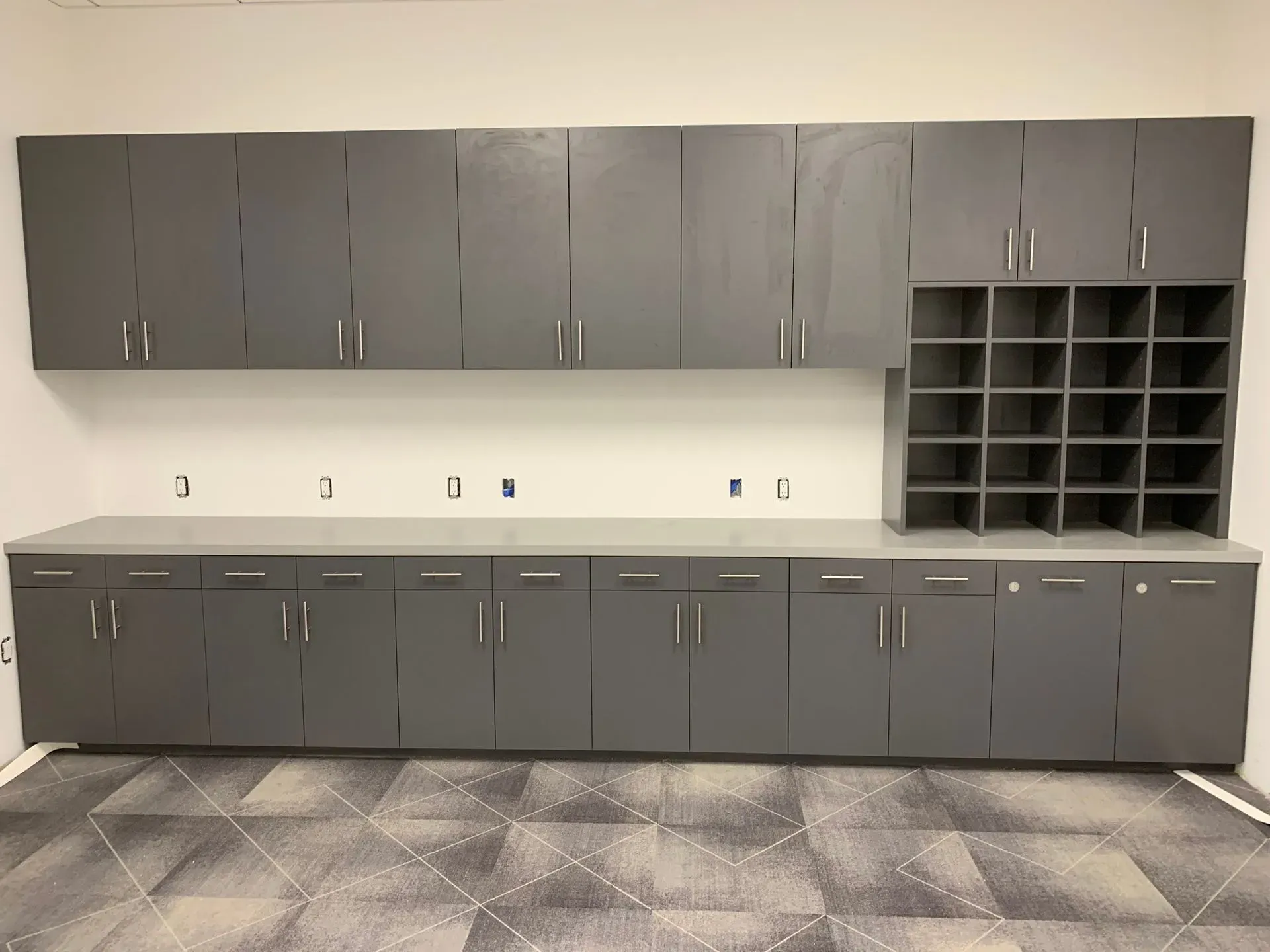 Gray cabinets and cubbies lining a room wall above and below a long countertop space.