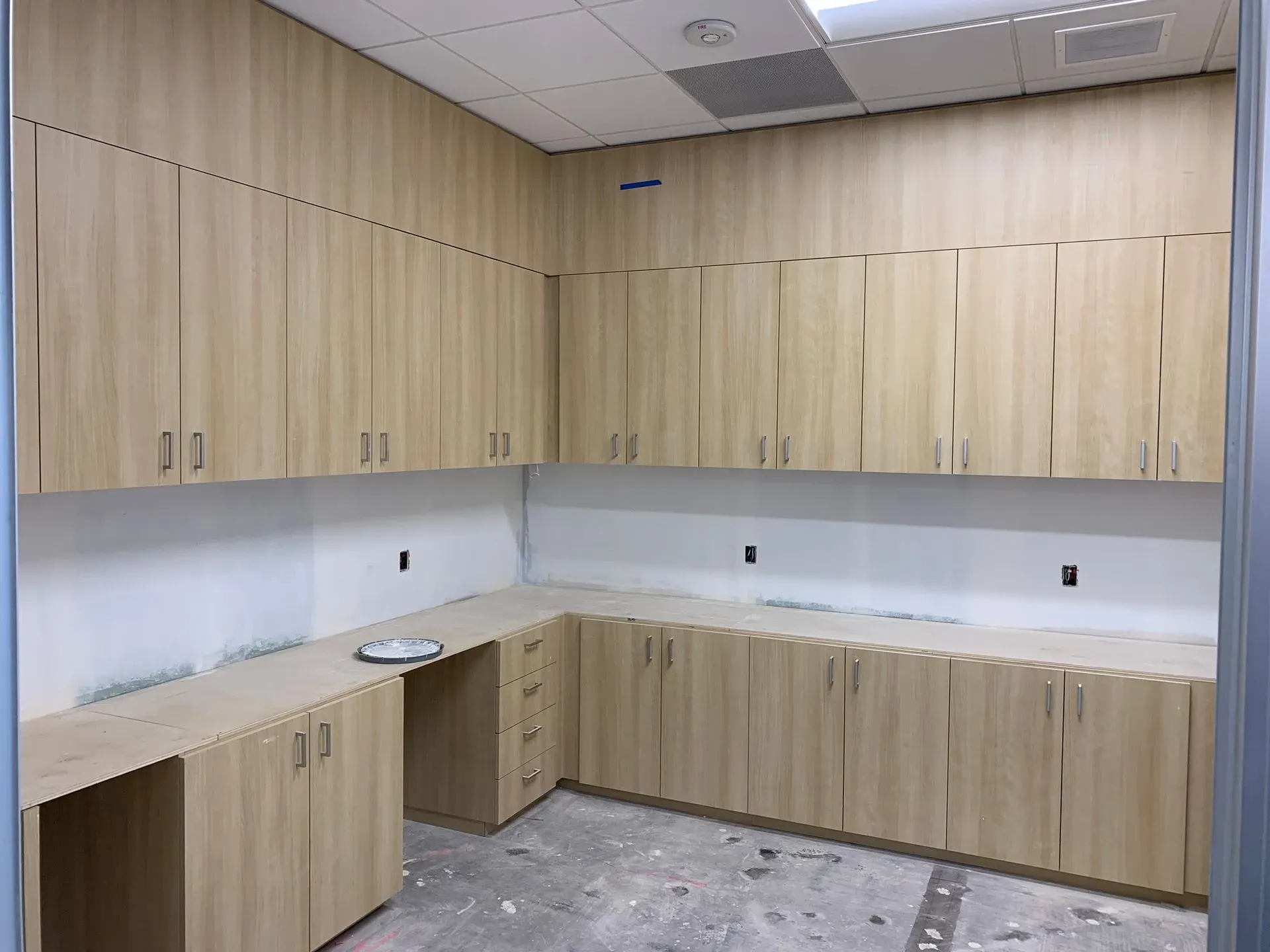 Empty office kitchenette with light wood cabinets and white countertops under bright ceiling lights