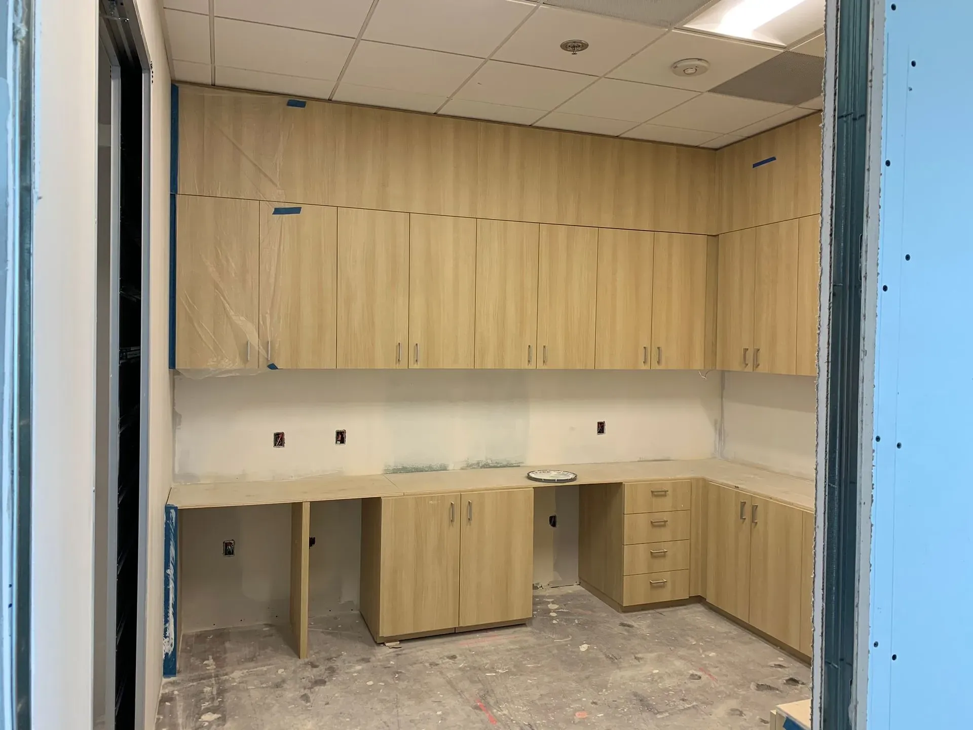 Empty office kitchenette with wood cabinets, white countertop, and a central cabinet island under construction.