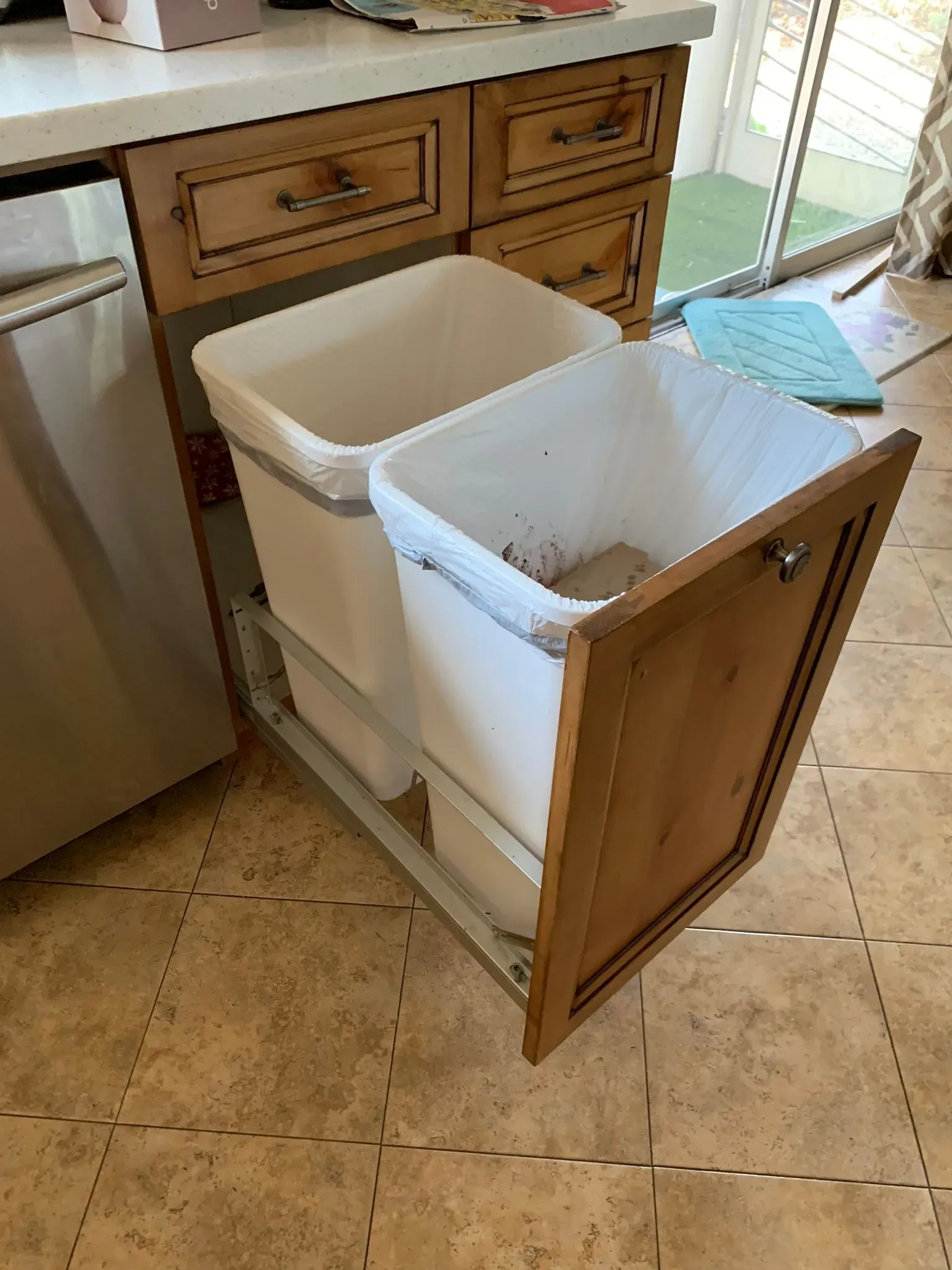 Pull-out kitchen trash bins in a wooden cabinet, with one bin door open on a tiled floor.