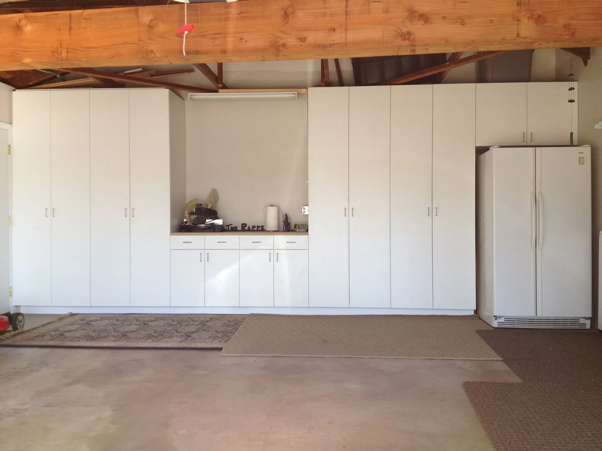 Garage wall with white storage cabinets, a small sink, and a refrigerator under exposed wooden rafters.