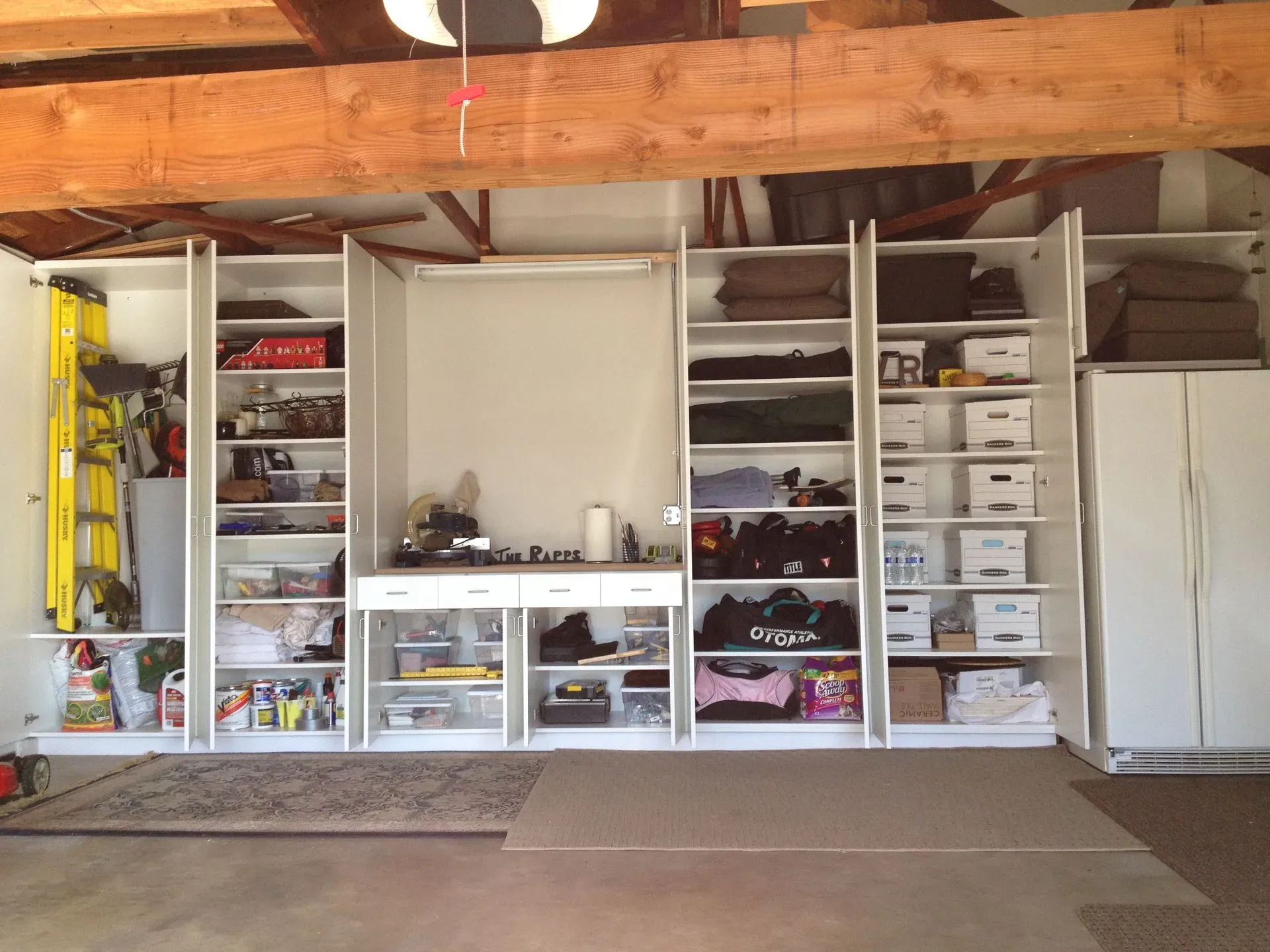 Garage storage wall with white shelving, plastic bins, tools, and a refrigerator under exposed wooden beams